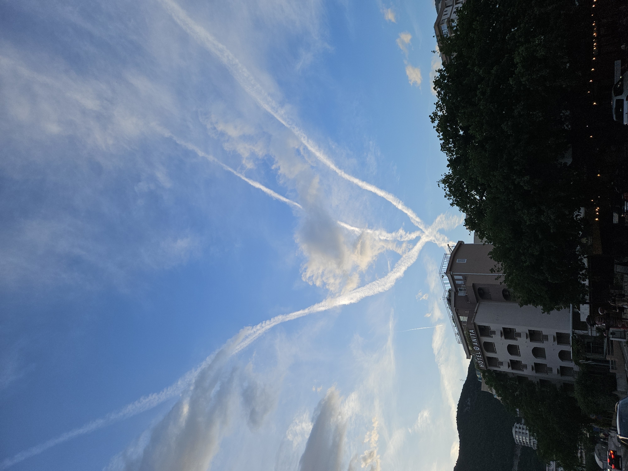 The image shows a daytime scene with a bright blue sky filled with contrails. In the lower right, there is a building labeled "HOTEL ANITA" surrounded by trees. There's a mountain visible behind the hotel. To the right of the building, there are trees and potentially city infrastructure, possibly a street with a car and street lights, but these are largely in shadow. The sky has many contrails creating an intricate network of lines and crosses. Some clouds are interspersed with the contrails. The image appears to be taken in the city of Unknown, Unknown.