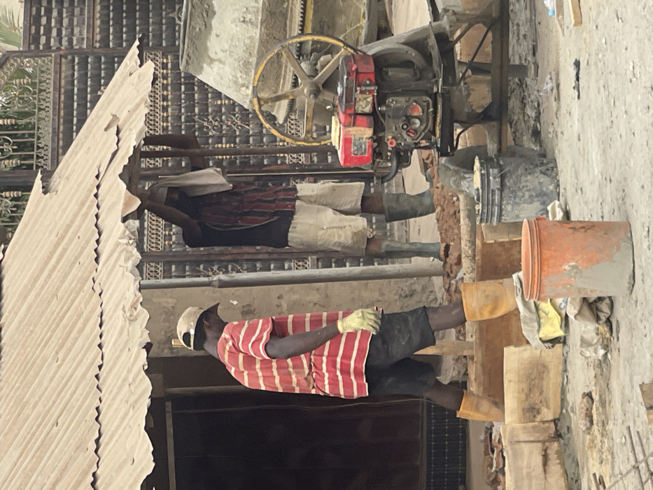Two men work on a construction site. The man in the foreground wears a red and white striped shirt, black shorts, and a white hat. He also wears yellow gloves. A second man, standing behind the first, wears a red and black plaid shirt. A cement mixer with a yellow wheel sits in the background. An orange bucket and other construction materials are visible. The ground appears to be dirt or concrete. Corrugated metal forms a partial roof.