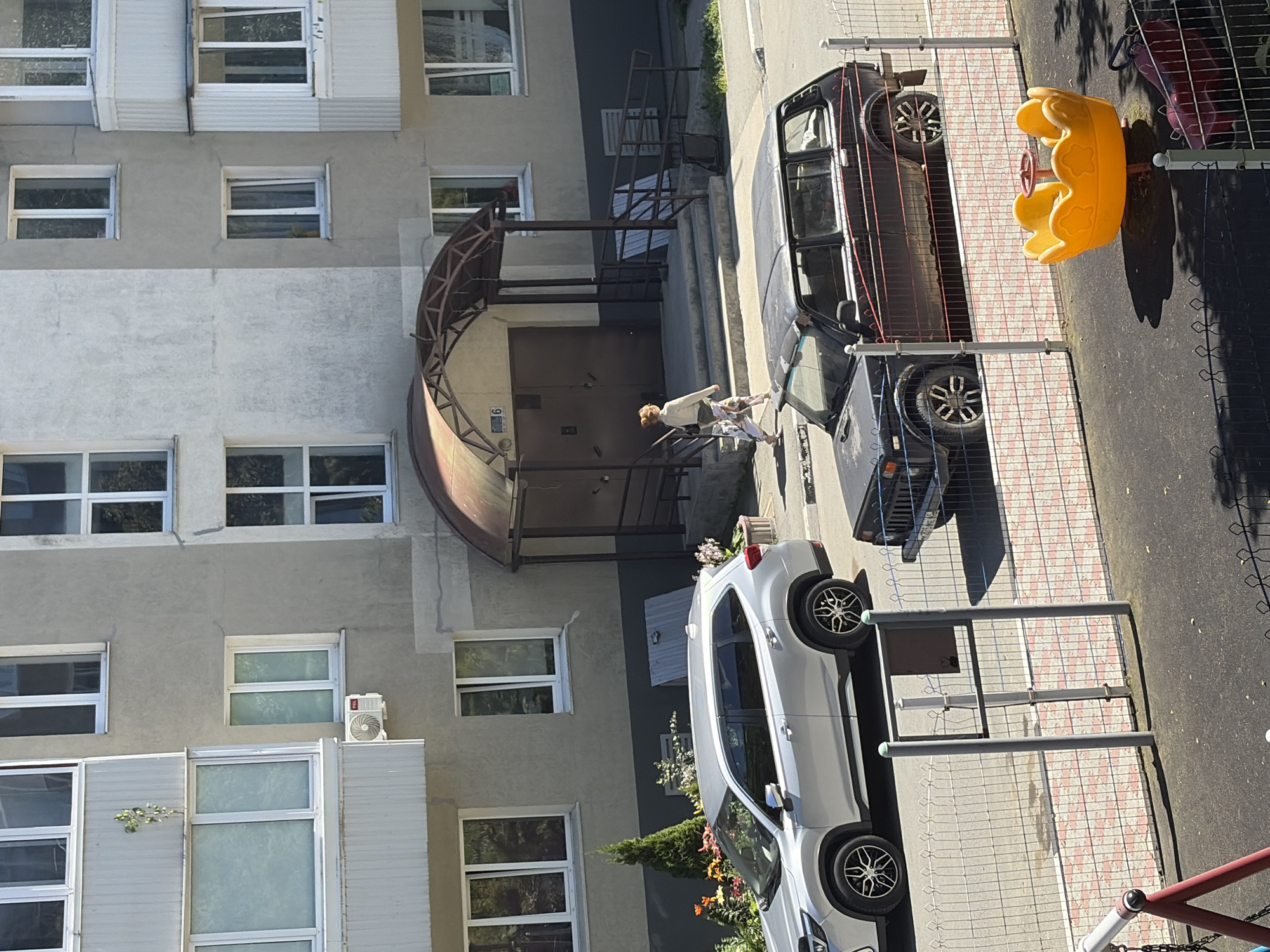 The image depicts a street scene in front of an apartment building, possibly in Korsakov, Russia. A silver SUV is parked on the street to the left, and a dark-colored older model hatchback is parked to its right, partially obscured by a fence. A woman with blonde hair is walking up the stairs towards the building entrance, which is covered by an arched canopy. The apartment building has multiple stories with visible windows. In the foreground, a playground area is visible with a yellow merry-go-round and a red slide. The area is paved and fenced.