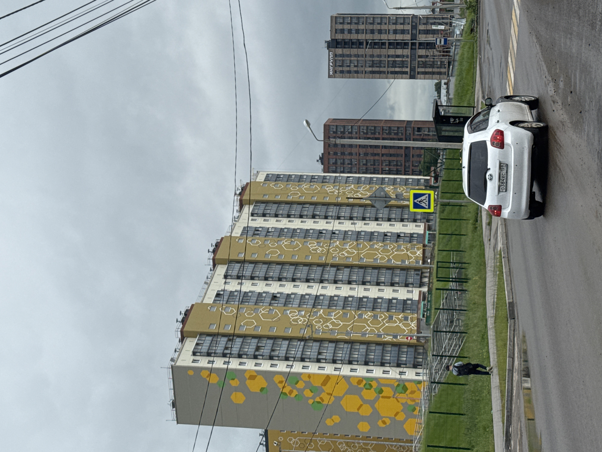 This image captures an urban street scene in Kondratovo, Russia, under an overcast sky. The foreground is dominated by a modern, multi-story residential building on the left, featuring a distinctive facade with large sections of yellow paint adorned with white and green hexagonal patterns. Further in the background, two taller, darker high-rise buildings are visible, one of which prominently displays the text "ИНГРУПП" (INGROUP) vertically on its side.
On the right side of the frame, an asphalt road features a clearly marked pedestrian crosswalk. A white Toyota car, with the Russian license plate "О384ХС 159," is positioned on the road near the crossing. A bright yellow pedestrian crossing sign stands prominently beside the road. Between the road and the main residential building, a grassy strip with a fence is visible, where a person in dark clothing, possibly wearing a face mask, is walking. A bus stop shelter is also partially visible further down the road.
Overhead, several power lines stretch across the light gray, cloudy sky. A street light is also part of the urban infrastructure visible in the scene. The road appears slightly wet, suggesting recent rain or damp conditions. The scene depicts a typical daytime setting in a developed residential area.