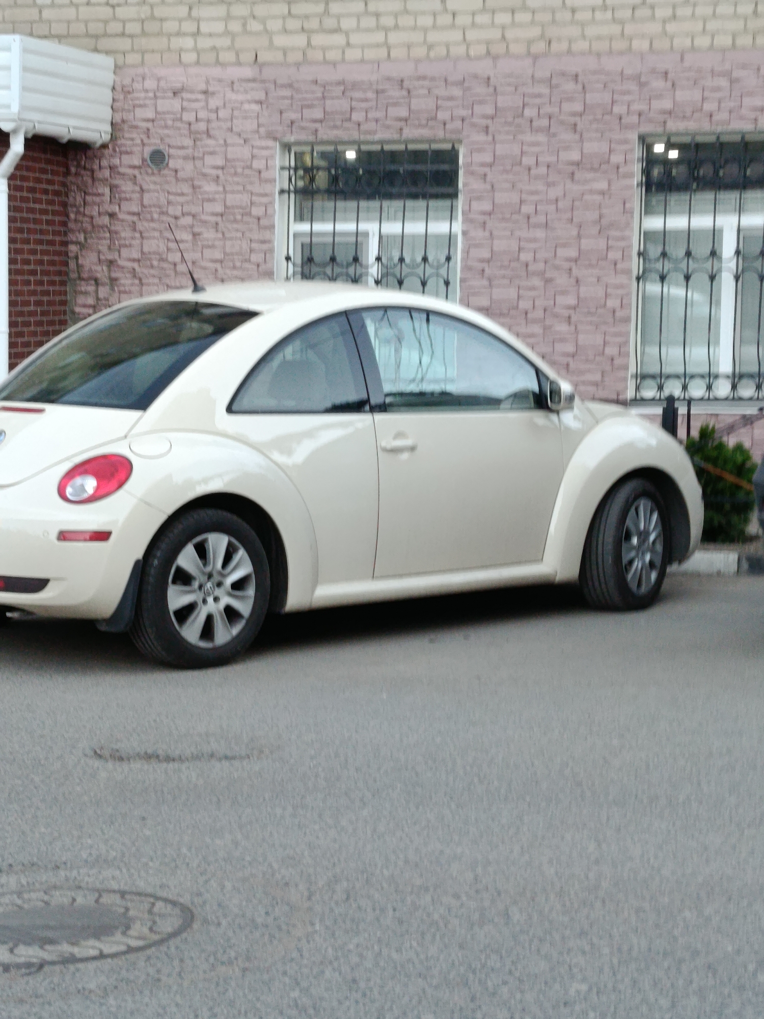A beige Volkswagen Beetle is parked on an asphalt surface in front of a brick building. The building has a pinkish-brown brick facade with two windows that have decorative black metal bars. To the left of the building is a section of red brick and a white air conditioning unit. The car is viewed from the side, with its rear half and part of its front visible. The car has tinted windows and silver alloy wheels. A small patch of green foliage is visible to the right of the car, behind the building. A manhole cover is visible on the asphalt in the lower left portion of the image. The scene appears to be a street or parking area in Saratov, Russia, based on the style of the building and the car's presence.