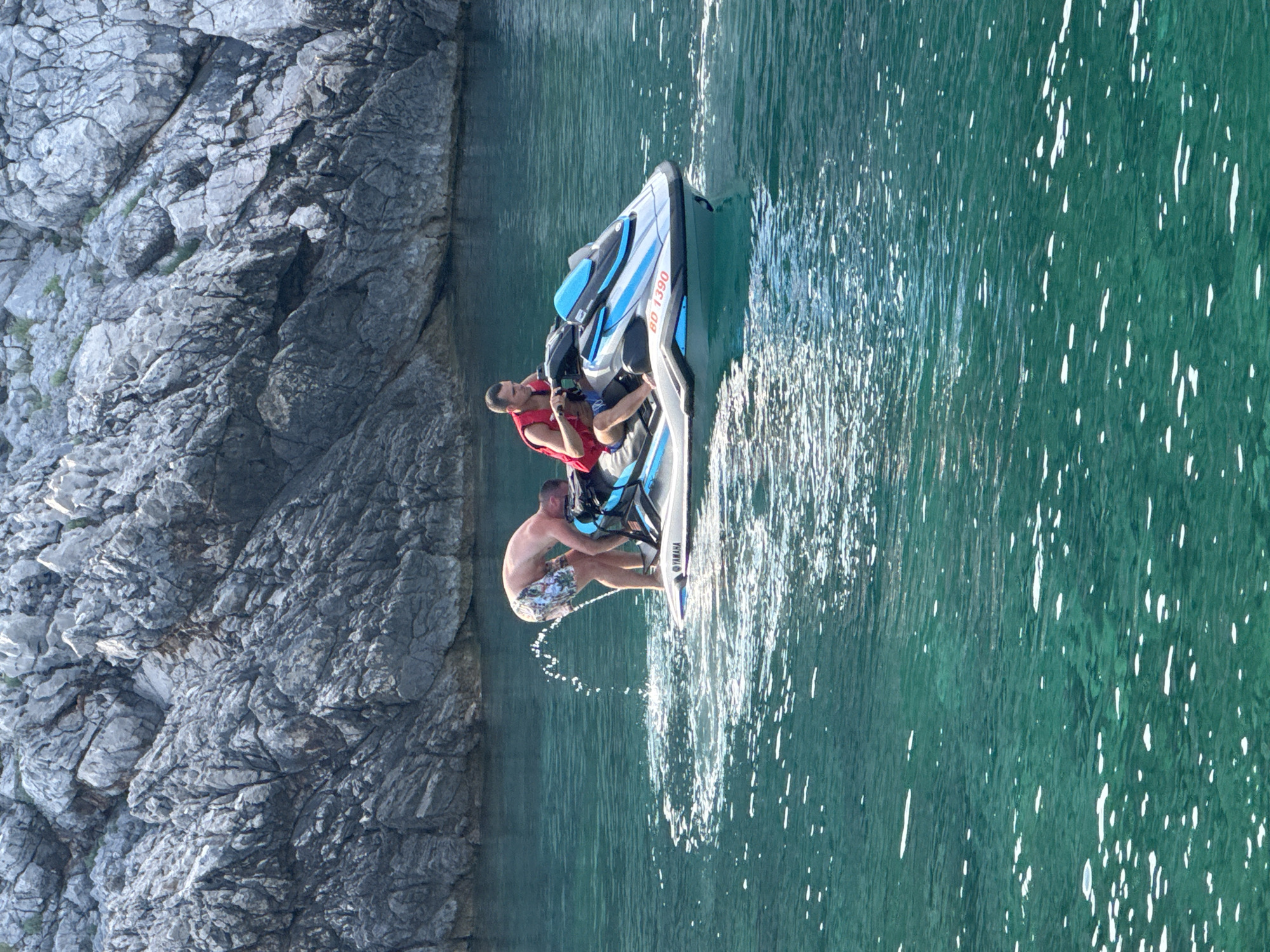 This media file shows two people on a jet ski on a sunny day, near a rocky coastline in Dubraljevina, Montenegro. The water is a clear, turquoise green, with sunlight reflecting off its surface. The rocky cliff face is gray and textured, showing signs of erosion.
The jet ski is white and blue, with the number "BD 1390" visible on its side, along with the "Yamaha" logo. One person, wearing a red life vest, is seated at the front of the jet ski, holding onto the handlebars. Another person, shirtless and wearing patterned swim shorts, is leaning back on the jet ski, with one leg extended and kicking water. They appear to be enjoying a recreational activity. The sunlight suggests it is daytime, likely afternoon given the shadows and the bright water. The overall mood is one of leisure and fun.
