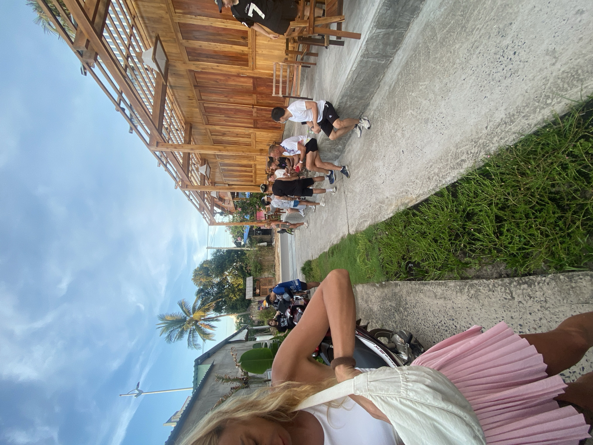 A group of individuals are gathered outside a wooden building with an open-air pergola structure. The building appears to be a cafe or restaurant, with wooden doors and a slatted roof. Several people are seated on a concrete ledge in front of the building, while others stand or are interacting with a motorcycle. The setting suggests a warm climate, with palm trees visible in the background. The individuals are casually dressed, indicating an informal gathering or waiting area. The scene captures a moment of daily life in what appears to be a semi-urban or resort environment in Phu Thuy Ward, Vietnam.