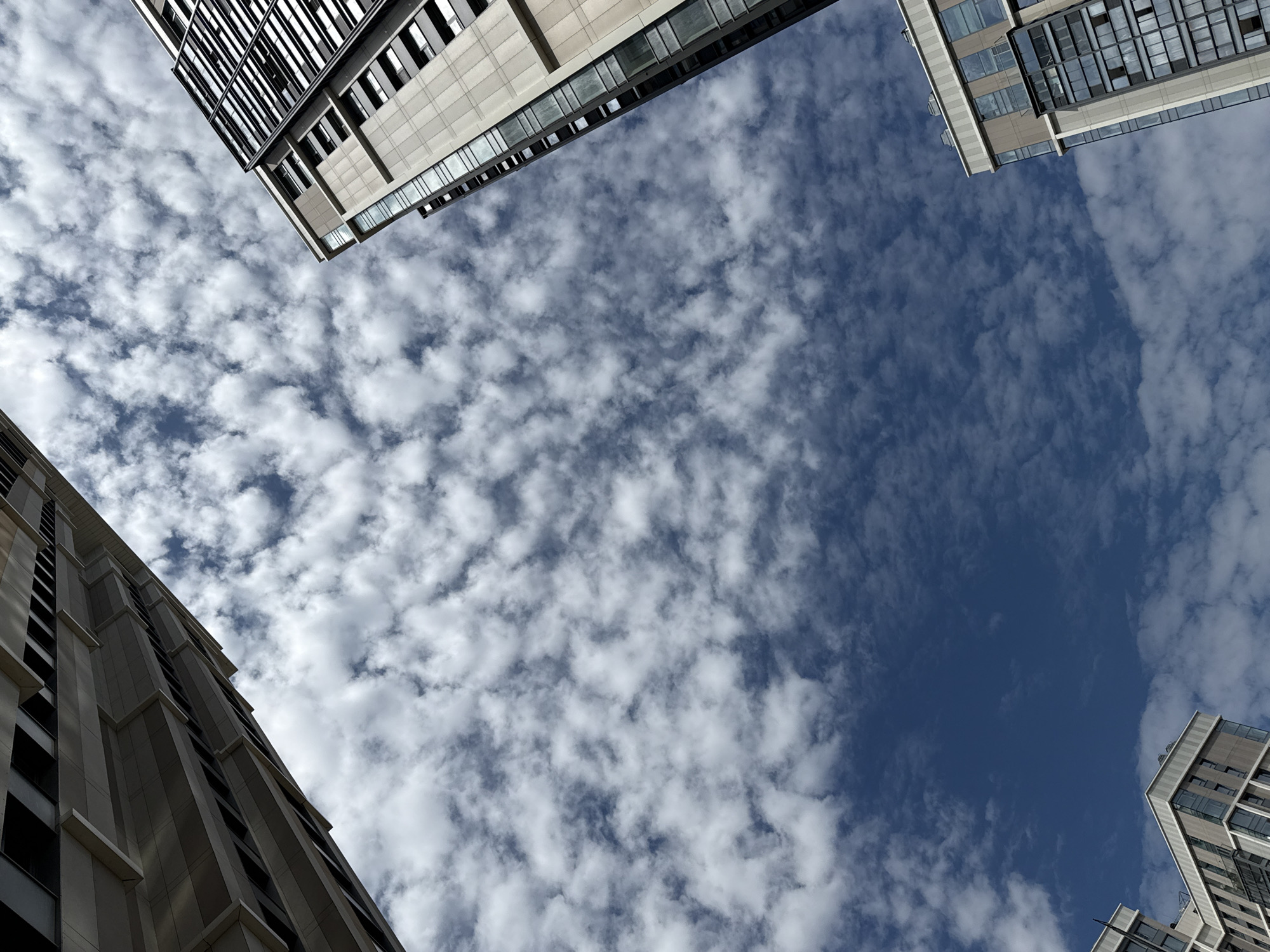 The image captures an upward-looking view of modern high-rise buildings against a sky filled with scattered white clouds. The architecture suggests a contemporary urban setting, likely in Khimki, Russia, given the context. The buildings are characterized by their geometric designs, large glass windows, and a palette of neutral tones, including beige, grey, and black. One building, prominently featured on the right, has a detailed facade with visible window panes arranged in a grid. Another building, partially visible on the upper left, shows a similar modern design. The sky is a vibrant blue, with fluffy clouds forming a dynamic pattern. There are no people or vehicles visible in the frame, focusing solely on the architectural elements and the natural environment.