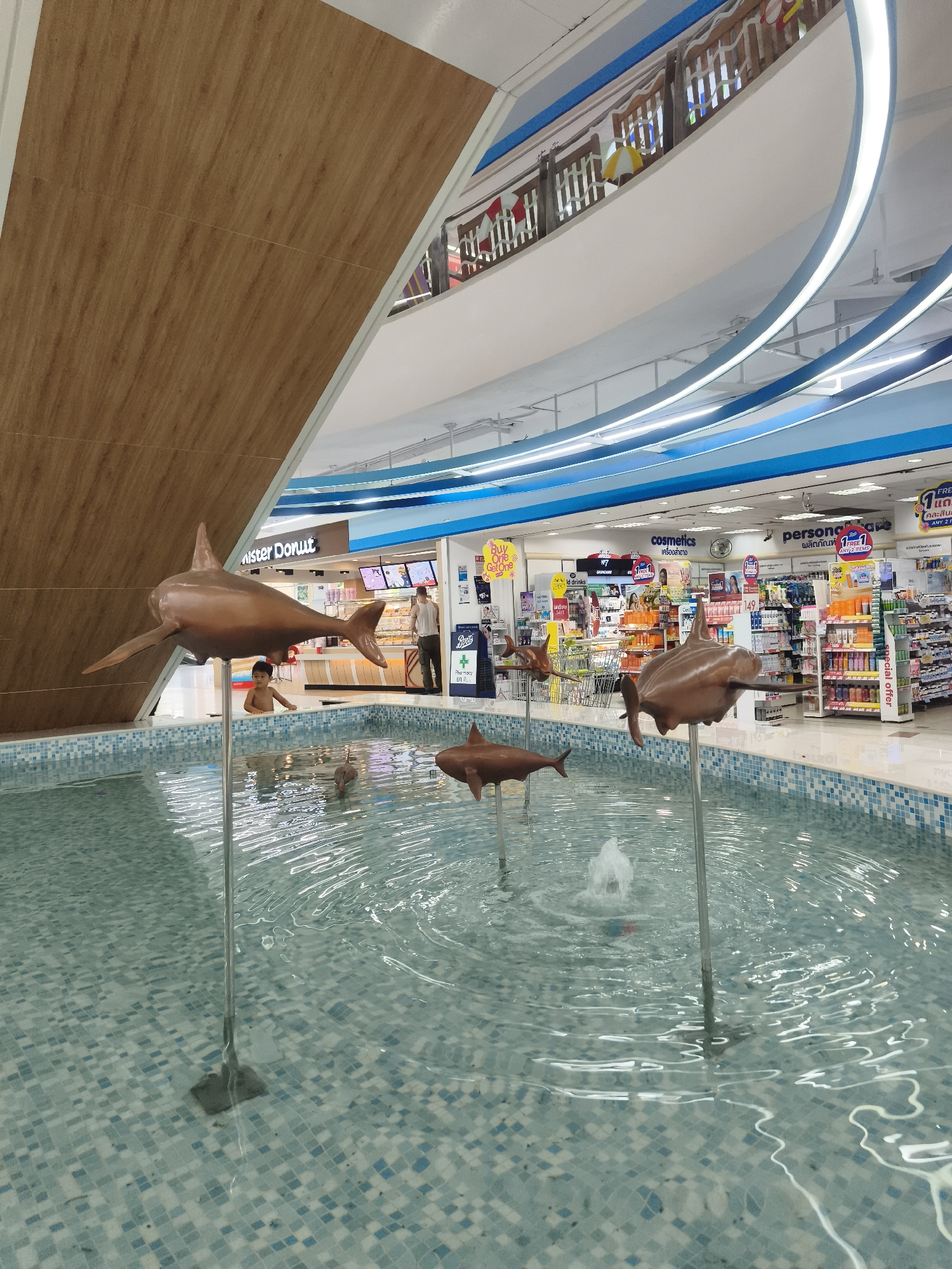 Here is a detailed description of the image:
**Scene:** The image shows an indoor space, likely a mall or shopping center, in บ้านบางรักษ์, Thailand. In the foreground is a tiled fountain filled with water. Within the fountain are several metal poles topped with copper-colored dolphin sculptures, creating a decorative water feature. A small fountain is in the middle.
**People:** A young child is seen at the edge of the fountain, facing forward into the fountain. An older man stands at the "Mister Donut" stand. Other individuals are vaguely visible within the stores in the background.
**Objects:** The background features a variety of retail stores, including a "Mister Donut" shop and a pharmacy with shelves stocked with various cosmetic and personal care products. Promotional signage for products like "cosmetics" and "personal care" is visible. There's a wooden facade to the left of the fountain. The shopping center's architecture is multi-tiered with open spaces and curved ceilings.
**Actions/Interactions:** The child is possibly playing near or in the water. The man at the donut shop is possibly serving customers.
**Environment:** The location is a well-lit, modern shopping environment. The overall ambiance suggests a casual and commercial setting in a Thai city.