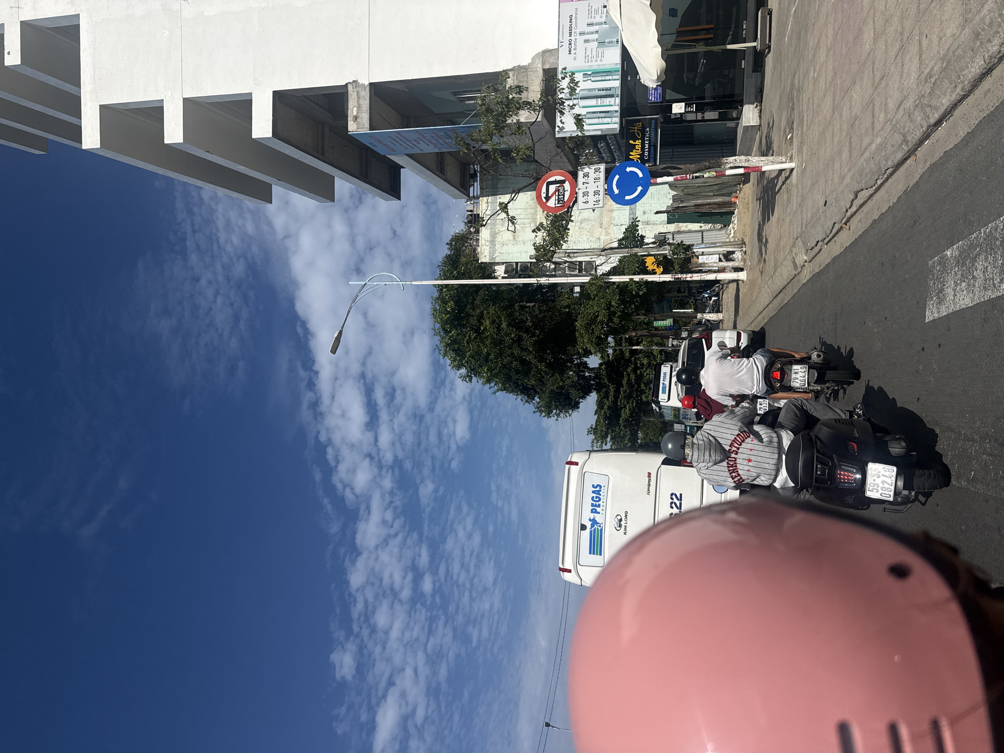 The image depicts a street scene in Nha Trang, Vietnam, on a sunny day with a bright blue sky and scattered clouds. The perspective is from behind a person wearing a pink helmet, facing forward towards traffic. In the foreground, a black scooter with a license plate reading "59-S3 082.48" is prominent, with a person in a grey hoodie with "ALENKO STUDIO" written on the back riding it. Another scooter is visible behind it. Further ahead, a white bus with "PEGAS TOURISTIK" and "KIM LONG" branding is stopped. A construction site with a modern, unfinished white building is on the right side of the street, featuring multiple levels and balconies. Traffic signs are visible on the sidewalk, including a circular red sign with a horizontal bar and a blue circular sign with three arrows indicating a roundabout. The sidewalk also has shops with signage, including "Minh Ha COSMETICS." Trees line the street, and the road surface shows a pedestrian crossing. The overall impression is of a busy urban environment with typical street activity.