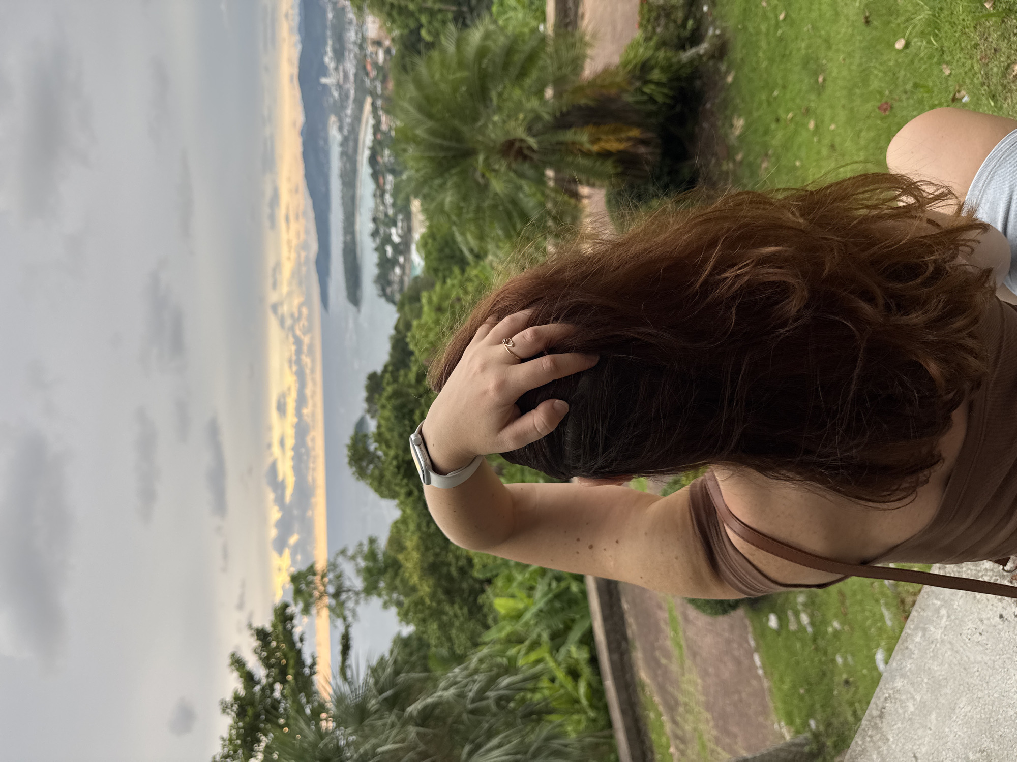 This is a vertical, medium shot of a person from behind, looking out at a scenic vista. The person has long, dark auburn hair that is slightly messy and pulled back with one hand. They are wearing a brown tank top and light-colored shorts. A smartwatch is visible on their left wrist. Their arm is extended up, resting on their head, and they are wearing a thin, delicate ring on their index finger.
The background is a lush, tropical landscape with palm trees and other dense foliage. Beyond the trees, a bay or coastline is visible, dotted with buildings and a harbor filled with boats. The sky is overcast with soft, golden light breaking through the clouds, suggesting either sunrise or sunset. The overall mood is peaceful and contemplative, with the person seemingly taking in the view. The location is Karon, Thailand. No text is visible in the image.