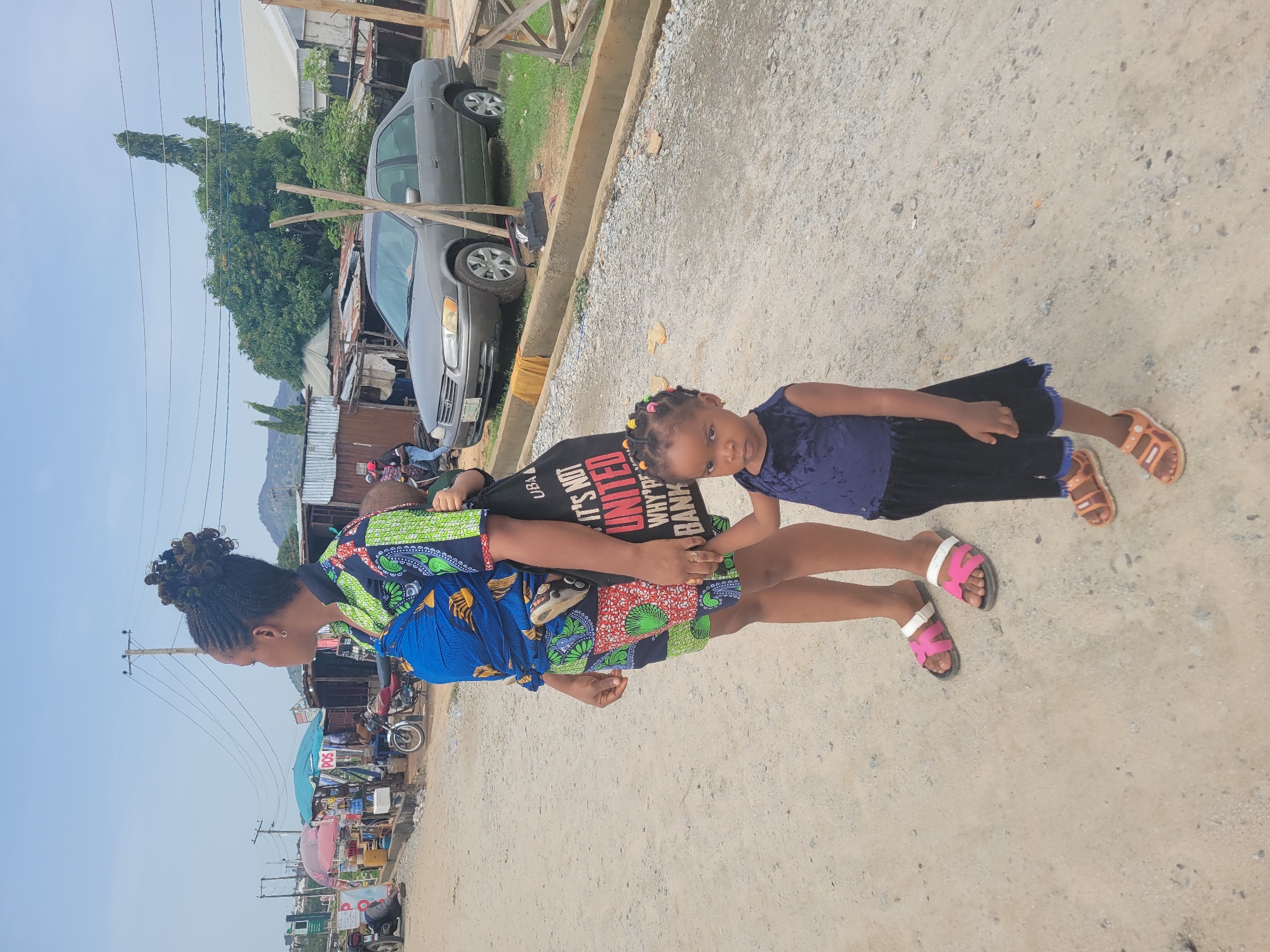 The image depicts a woman and a young girl standing on a dirt road in Kwamba, Nigeria. The woman, with braided hair, is wearing a colorful patterned dress and pink sandals. She is carrying a child on her back, and the child wears a black t-shirt with white writing. The young girl standing beside her is wearing a dark blue dress and brown sandals. In the background, there is a car parked next to a curb and multiple street vendors.