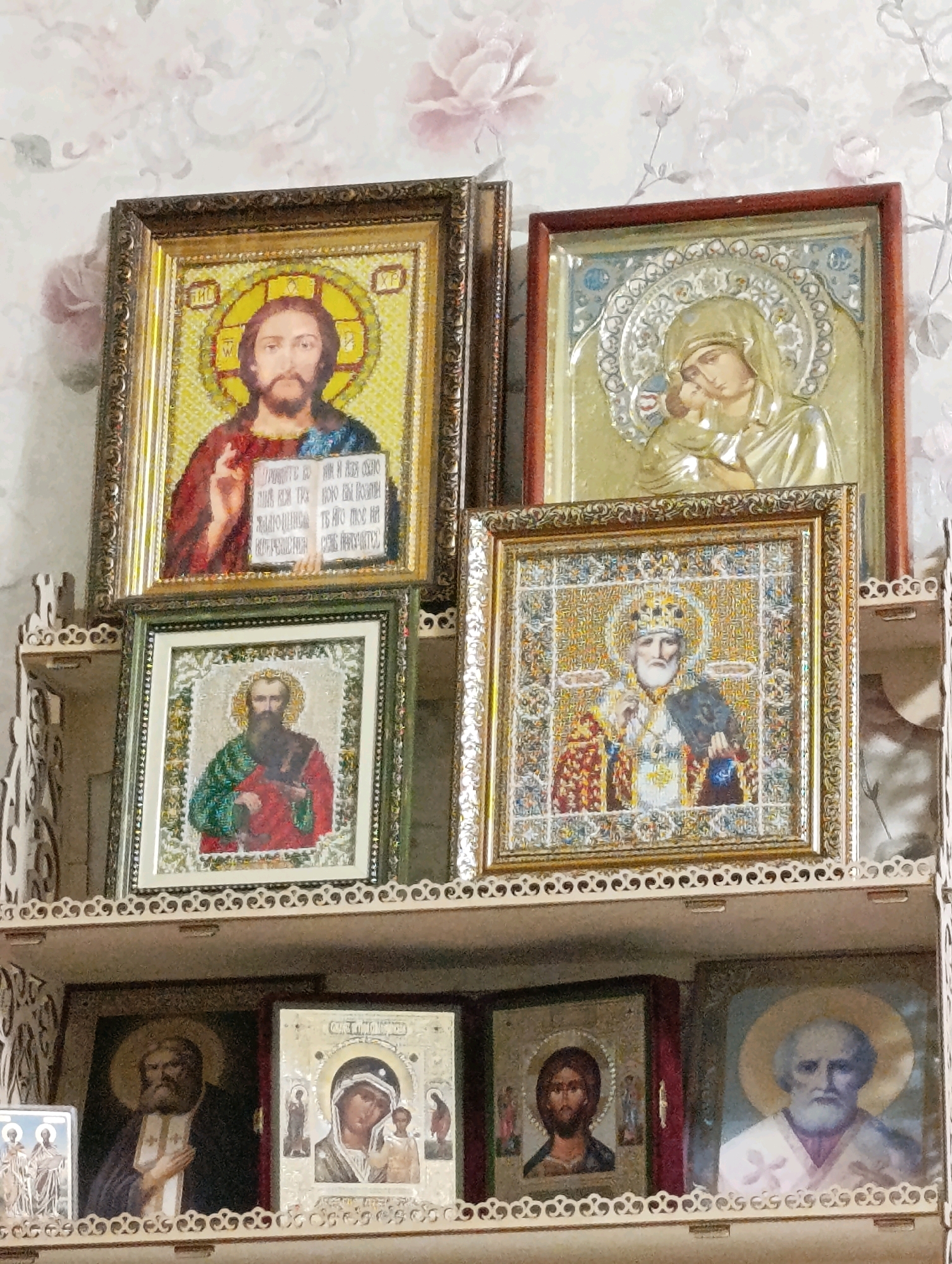 The image shows a collection of Orthodox Christian icons displayed on a tiered shelf against a floral wallpaper background. The icons vary in size and style, with some appearing to be painted and others possibly made with mosaics or embroidery.
In the top row, on the left, is an icon of Jesus Christ, depicted with a halo and holding an open book. To its right is an icon of the Virgin Mary holding the infant Jesus, also with halos.
The middle row features two icons. On the left, a smaller icon depicts a bearded saint in red and green robes. On the right, a larger, ornate icon shows a saint wearing a crown and bishop's vestments, holding a book.
The bottom row displays several more icons on a lower shelf. From left to right, the visible icons include: a depiction of multiple figures, an icon of Jesus Christ, and an icon of a bearded man in religious attire with a halo. The shelf itself has decorative carvings. The overall impression is of a personal devotional space.
