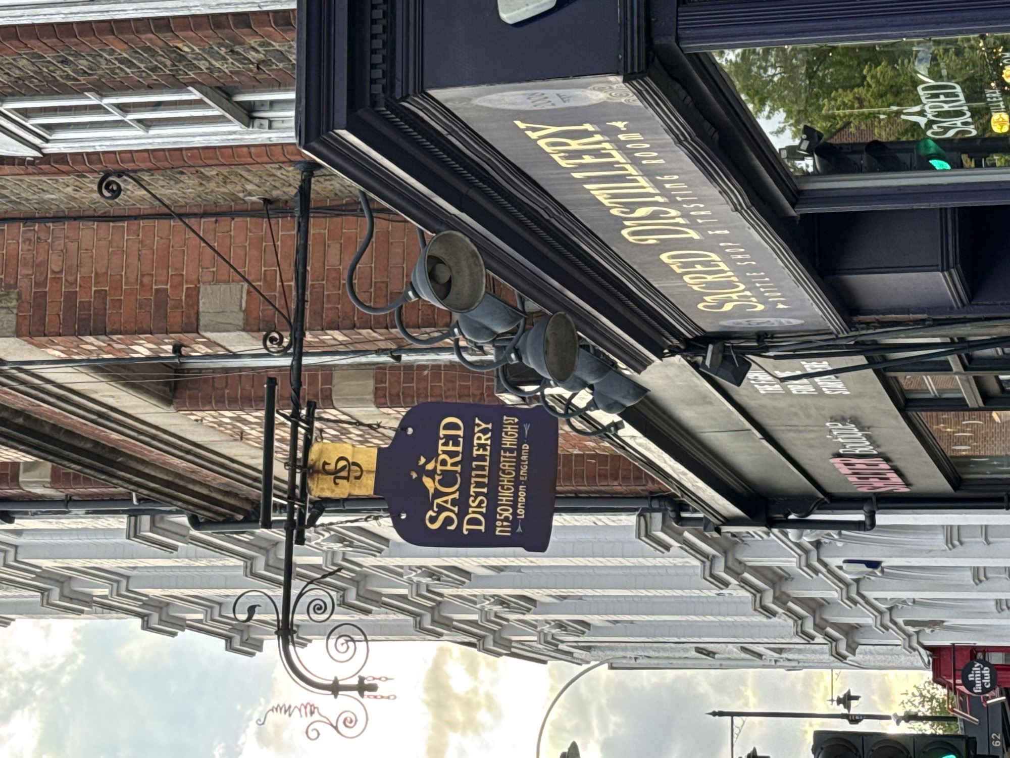Here is a description of the image:
The image shows a street scene in London, United Kingdom, featuring the storefront of "Sacred Distillery." The camera angle is looking up at the signs and storefronts. The "Sacred Distillery" sign hangs perpendicular to the building, displaying the name and address (No. 50 Highgate High St, London England) in gold lettering on a dark background. Above this is a larger, horizontal sign also indicating "Sacred Distillery - Bottle Shop & Tasting Room" in gold. There are three gray spotlight-like lights directed towards the sign.
The surrounding architecture consists of brick and white-painted structures. A traffic light with a green light is visible in the background. A red and white “The Family Club” sign can be seen in the lower right corner. The sky is visible with some cloud cover.