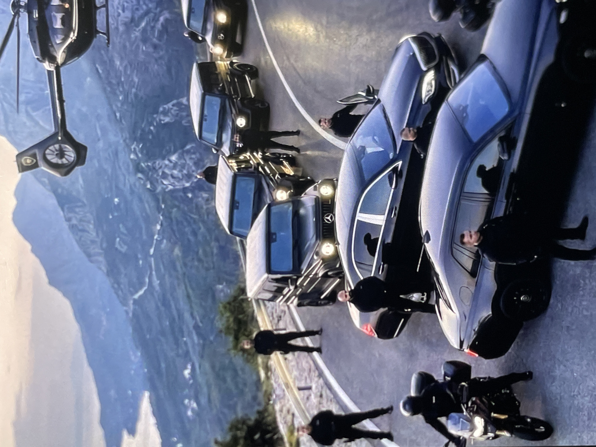 An aerial shot captures a motorcade on a winding mountain road. A helicopter hovers above, while several black SUVs and luxury sedans are stopped on the asphalt. Numerous individuals, dressed in dark tactical gear, stand guard near the vehicles, some appearing to be security personnel. The rugged, mountainous terrain is blanketed in a soft haze, with glimpses of a distant coastline and body of water. The scene suggests a high-profile transport or security operation in a remote, scenic location.