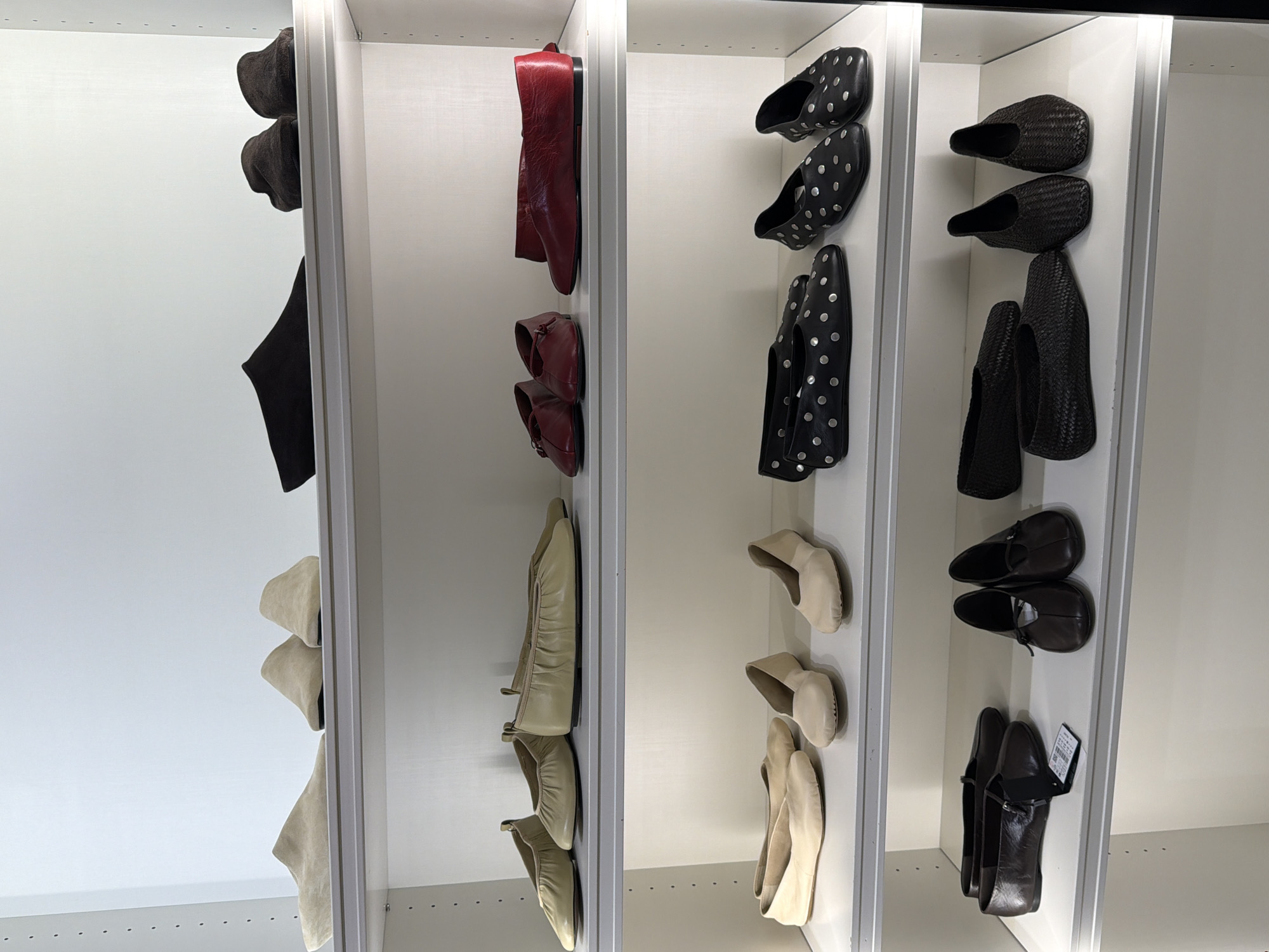 The image displays a closet with multiple shelves organized to store shoes. The closet appears to be part of a retail store or a well-organized personal wardrobe. The lighting suggests it is daytime, with artificial light illuminating the interior of the closet.
On the leftmost shelf, there are several dark-colored, soft-brimmed hats, stacked on top of each other. Below the hats, a pair of black boots are displayed, with one boot positioned upright and the other lying on its side.
The second shelf from the left holds a pair of red leather ankle boots. Below these, a pair of darker red leather shoes with buckles are visible.
The third shelf features black slip-on shoes adorned with silver polka dots. There are two pairs of these shoes, with the top pair facing forward and the bottom pair positioned slightly behind.
On the fourth shelf from the left, there are two pairs of dark brown woven slip-on shoes, stacked one above the other. Below these, two pairs of dark brown leather loafers are displayed, one of which has a tag attached.
The fifth shelf contains two pairs of light beige ballet flats.
The shelf furthest to the right showcases several pairs of light beige shoes. The top two pairs are slip-on shoes, with one pair facing forward and the other angled. Below these, two pairs of ballet flats are displayed.
The overall scene is neat and orderly, showcasing a variety of footwear organized by style and color. There is no indication of people or any specific activity taking place beyond the display of the shoes. The setting is an indoor closet, likely in a location with organized storage solutions. No visible text beyond a shoe tag on the bottom right shelf.