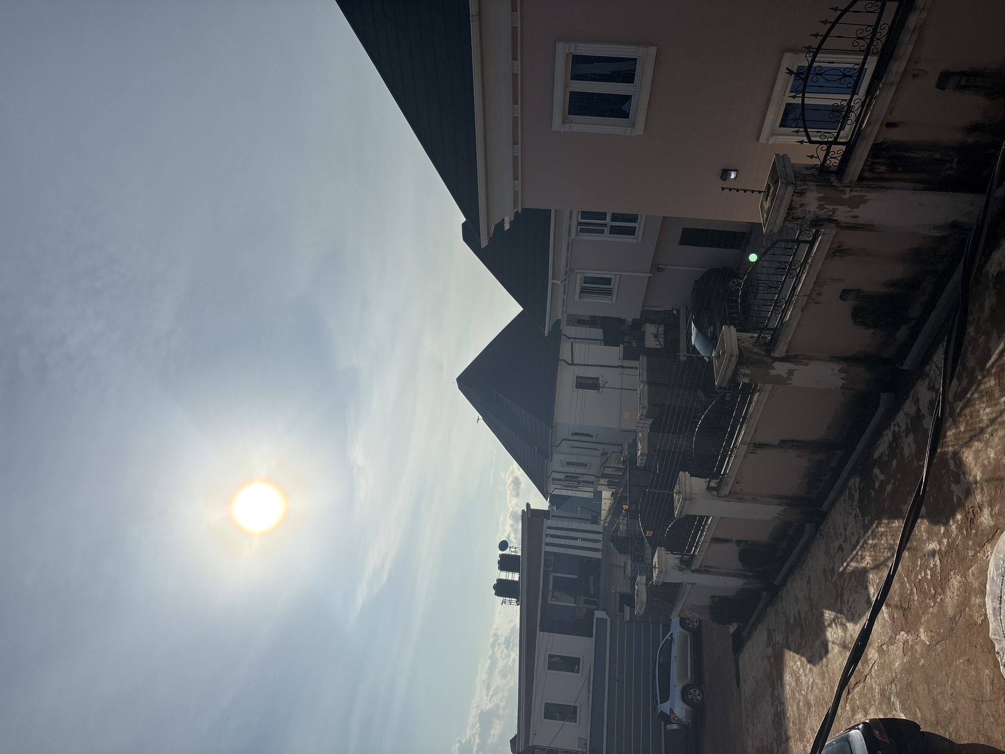 This is an exterior, eye-level shot of a residential area in Obosi, Nigeria, under a bright, partly cloudy sky. The sun is high and casting strong shadows. Several modern houses with dark-tiled roofs and pale exteriors are visible. One house on the left has a white and red facade with prominent water tanks on its roof, and a silver SUV parked in its driveway. Adjacent to it, another white house features a pointed dark roof. To the right, a blush-pink house has a similar dark roof and large windows. A light pink stucco wall topped with ornate black metal fencing encloses the properties. The ground is a mixture of reddish-brown soil and paved areas. The overall impression is one of contemporary, upscale housing in a sunny, urban or semi-urban environment.