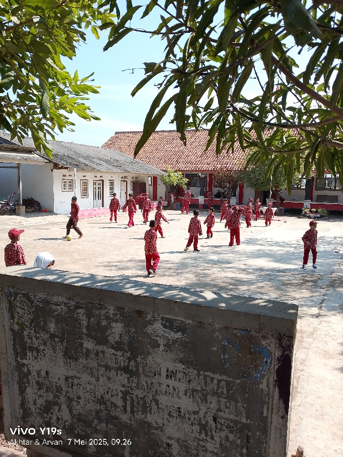The image shows a schoolyard in Kecamatan Haurgeulis, Indonesia (latitude: -6.419, longitude: 107.972). A group of children, all wearing red shirts and pants, are playing in a large, paved courtyard surrounded by buildings. There are three main buildings with tiled roofs, one of which appears to be a school building with windows. The courtyard is partially shaded by a large tree in the foreground, whose branches are reaching out towards the buildings. The children are engaged in various activities. Some are running around, while others are kicking a soccer ball, which is visible in the center of the image. The sun is shining, and there is a cast shadow from the tree in the foreground. The ground is dusty and covered with some leaves and pebbles. A large, weathered concrete wall with faded writing on its surface is in the foreground. The wall is covered in a thin layer of dust and debris. The writing on the wall is obscured but is partially visible. The image appears to have been captured by a cell phone camera.