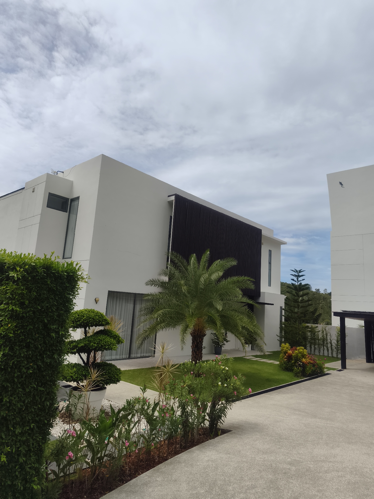 This image captures the exterior of a modern, multi-story white building, likely a villa or part of a resort, under an overcast sky. The architecture features clean lines, large windows, and a dark, vertically slatted facade on a section of the main structure.
In the foreground, a meticulously maintained garden and concrete pathways lead up to the building. Lush green landscaping includes a tall, manicured hedge on the left, a large potted bonsai-style tree, and various flowering plants with pink and white blooms lining the path. A prominent palm tree stands centrally in the garden, alongside a vibrant green lawn.
To the right, a second white building of similar design is visible, featuring a distinct blue lizard or gecko emblem on its upper facade. A dark, open-sided structure, possibly a car shelter or entryway, is also present on the right. In the background, rolling green hills covered in dense foliage extend under a sky filled with scattered white clouds, suggesting a tropical location.
The scene is set during the daytime, with soft, diffused light from the cloudy conditions. No people or activities are visible, emphasizing the tranquility and pristine nature of the property. The overall impression is one of contemporary elegance and meticulous upkeep within a natural, serene environment, consistent with a high-end residential or hospitality setting in Baan Tai, Thailand.