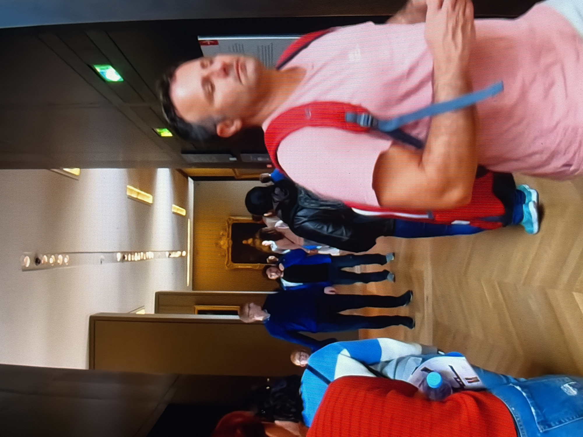 The image depicts an indoor scene, likely a museum or gallery, with a man in a pink t-shirt and red backpack in the foreground, facing right and slightly away from the camera. He has short, dark hair and appears to be in motion. Behind him, several other individuals are visible, including a man in a blue sweater and glasses, and people in casual attire. The environment features wooden flooring, high ceilings with lighting fixtures, and artwork on the walls, including a large framed painting in the background. Green emergency exit signs are visible on a dark wall to the right. The lighting suggests an interior setting with artificial illumination.