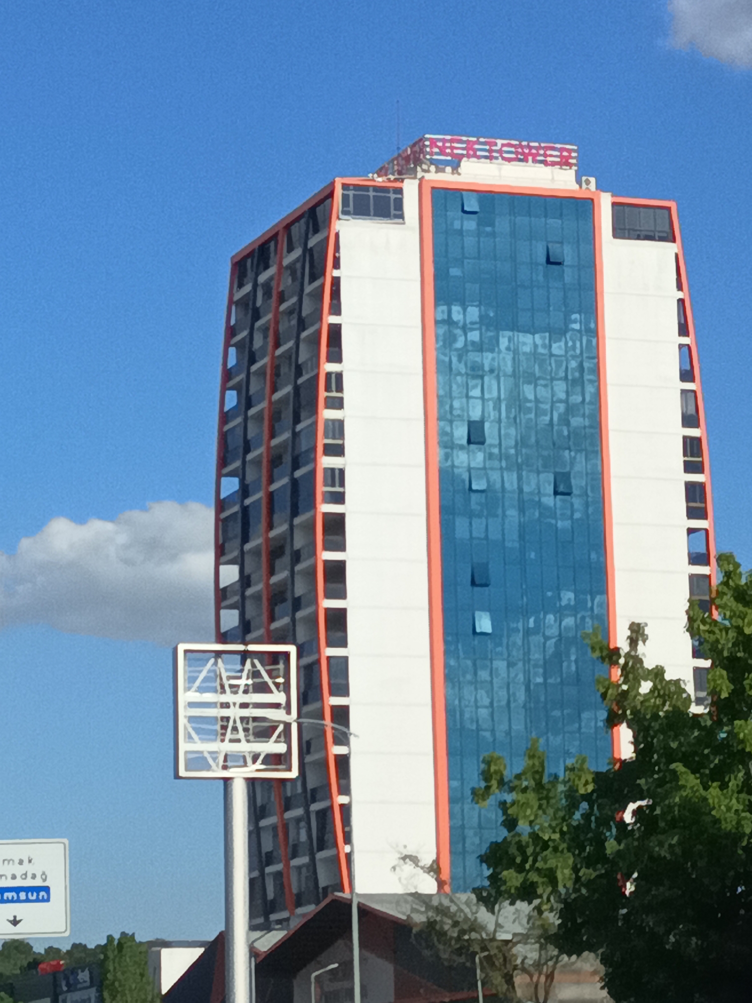 This media file shows a tall, modern building with an orange frame and white walls, featuring large blue glass windows. The building appears to be an office building or hotel. It's a sunny day with a bright blue sky and some light clouds. In the foreground, there's a street sign with Turkish text. The sign appears to be a directional sign, pointing downwards with an arrow. To the right of the building, there are some green trees. The building has a logo or text at the top, which reads "BİLFEN TOWERS" in red and white letters. The overall scene suggests an urban environment in Turkey, likely during daytime. There are no people visible in the frame.