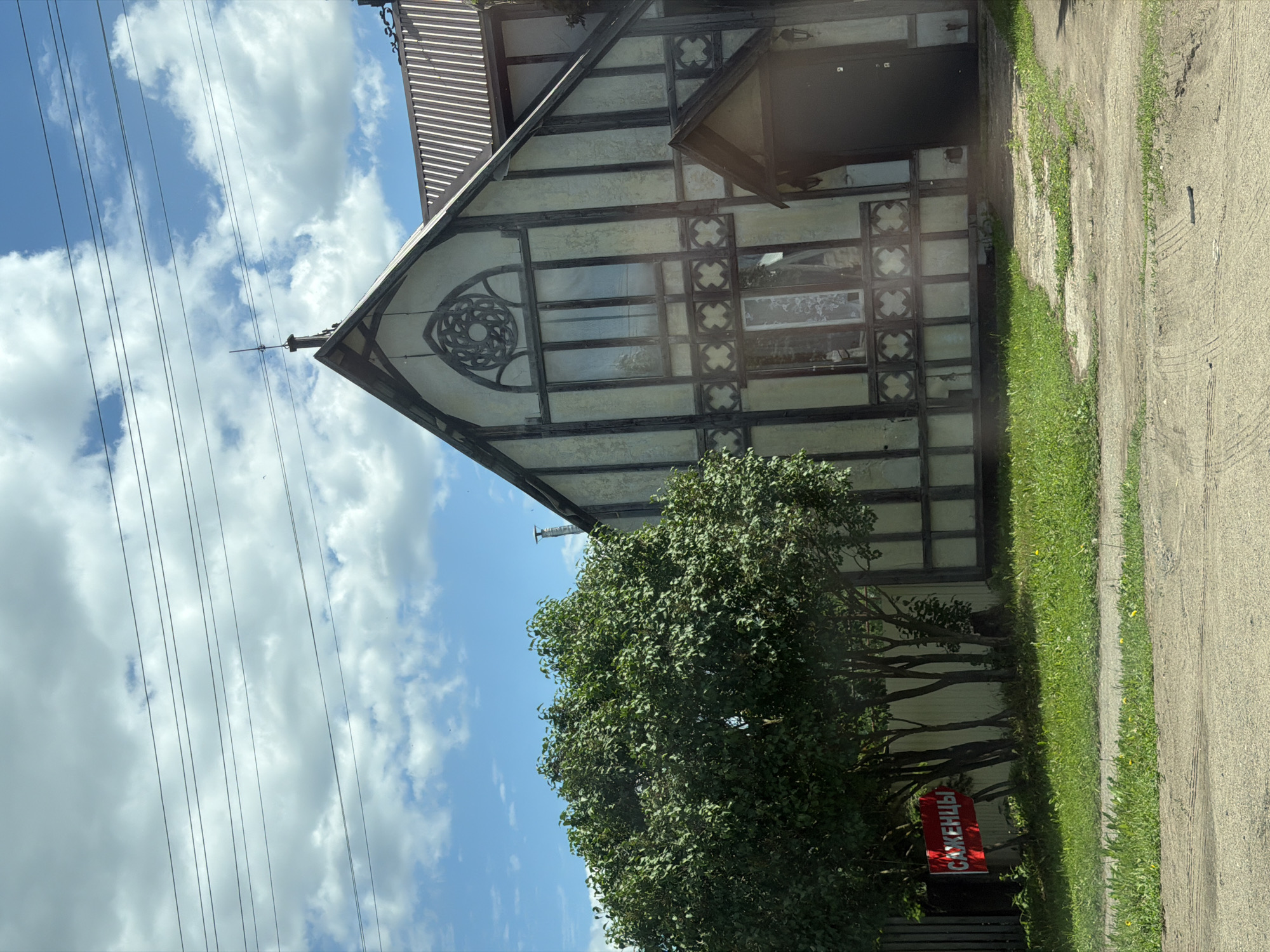 The image depicts a two-story residential structure of distinct Tudor Revival architectural style, featuring exposed dark timber framing over light-colored infill panels. The house is situated in what appears to be a semi-rural or suburban environment, potentially in Кварса, Russia. The front facade presents a prominent gable end, centrally adorned with a large, intricately carved round window (resembling a rose window) above multiple vertical window panes. Below these, a decorative horizontal band of quatrefoil-like patterns is incorporated into the timberwork. A recessed entrance with a small gabled porch and a dark door is visible on the lower right side of the facade, accompanied by a wall lantern. The house is topped with a steeply pitched roof, transitioning to a corrugated metal roof section on the right, from which a metal chimney or vent pipe emerges. A small finial crowns the main gable's apex.
To the left of the house, a large deciduous tree with dense green foliage partially obscures the building. Beneath this tree, a red rectangular sign with white Cyrillic text, "САЖЕНЦЫ" (SAPLINGS), is affixed to a dark fence, pointing towards the right. The ground in the foreground consists of a dirt or gravel path/road, transitioning into a strip of green grass containing some yellow flora, possibly dandelions. A lighter-colored, possibly concrete path leads from the foreground towards the house's entrance.
The sky above is bright blue with scattered, fluffy white cumulus clouds. Multiple parallel utility lines traverse the upper portion of the frame, horizontally spanning across the sky and above the house. No individuals are present within the scene. The lighting suggests a clear, sunny day, likely during late morning or early afternoon hours.