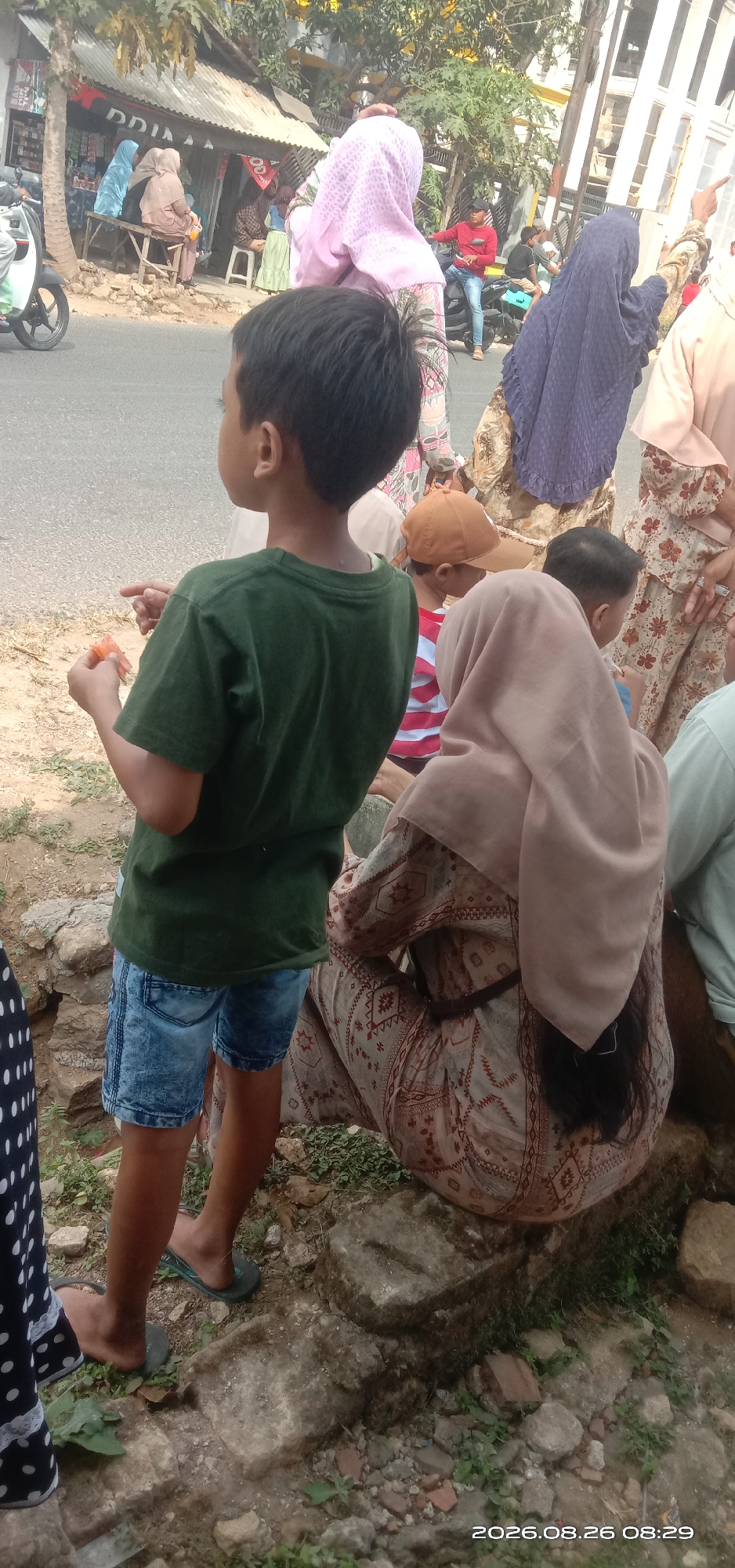 A group of people are gathered outdoors in a street scene, likely in a residential area or near a marketplace in Pakong, Indonesia. In the foreground, a young boy in a green t-shirt and denim shorts stands facing away from the camera, holding a piece of orange food. To his right, a woman wearing a light brown hijab and a patterned dress is seated. Behind them, other individuals are visible, some wearing hijabs and traditional clothing. A motorcycle is parked on the street in the background, and a small building with a corrugated metal roof is also present. The ground is a mix of dirt, gravel, and some scattered stones. The overall atmosphere appears to be casual and communal.