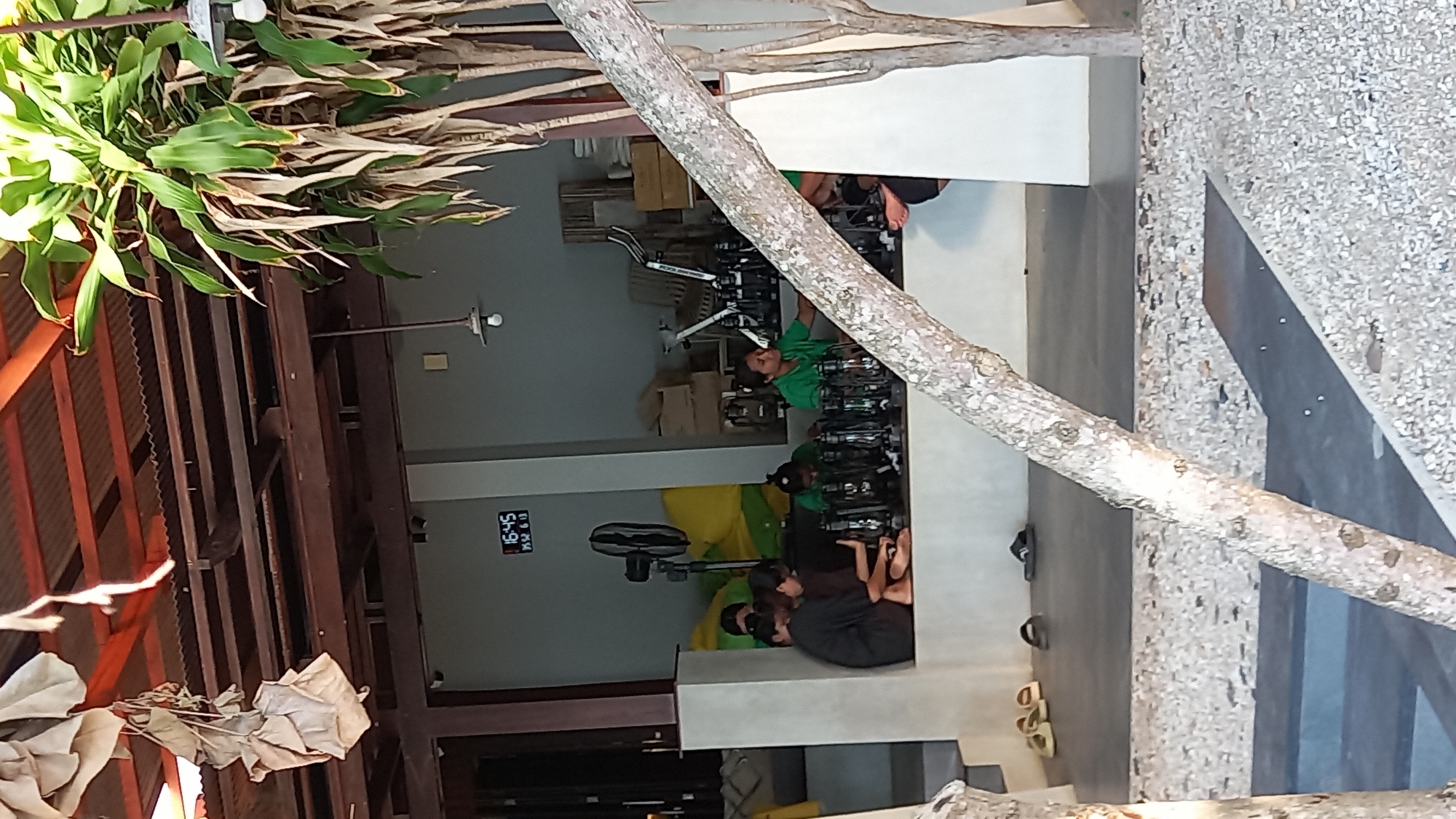 The image depicts an outdoor seating area with several people gathered around a counter, possibly engaging in a workshop or event. The environment appears to be a cafe or restaurant with a rustic, natural aesthetic. The roof is made of corrugated metal supported by wooden beams, and there are hanging decorations made of fabric. In the background, a digital clock displays "16:45" and "19.02.13", suggesting the time and date. Several individuals are seated, some interacting with what appear to be portable lamps arranged on the counter. One person in a green shirt is facing the camera, while others are looking down or away. To the left, a large black fan is visible. The ground is paved with a textured surface of small stones. In the foreground, a tree trunk branches across the frame, partially obscuring the view of the seating area. The overall impression is a relaxed, daytime gathering in a tropical or semi-tropical setting. The location is identified as Koh Phangan, Thailand, suggesting a beachside or island resort atmosphere.