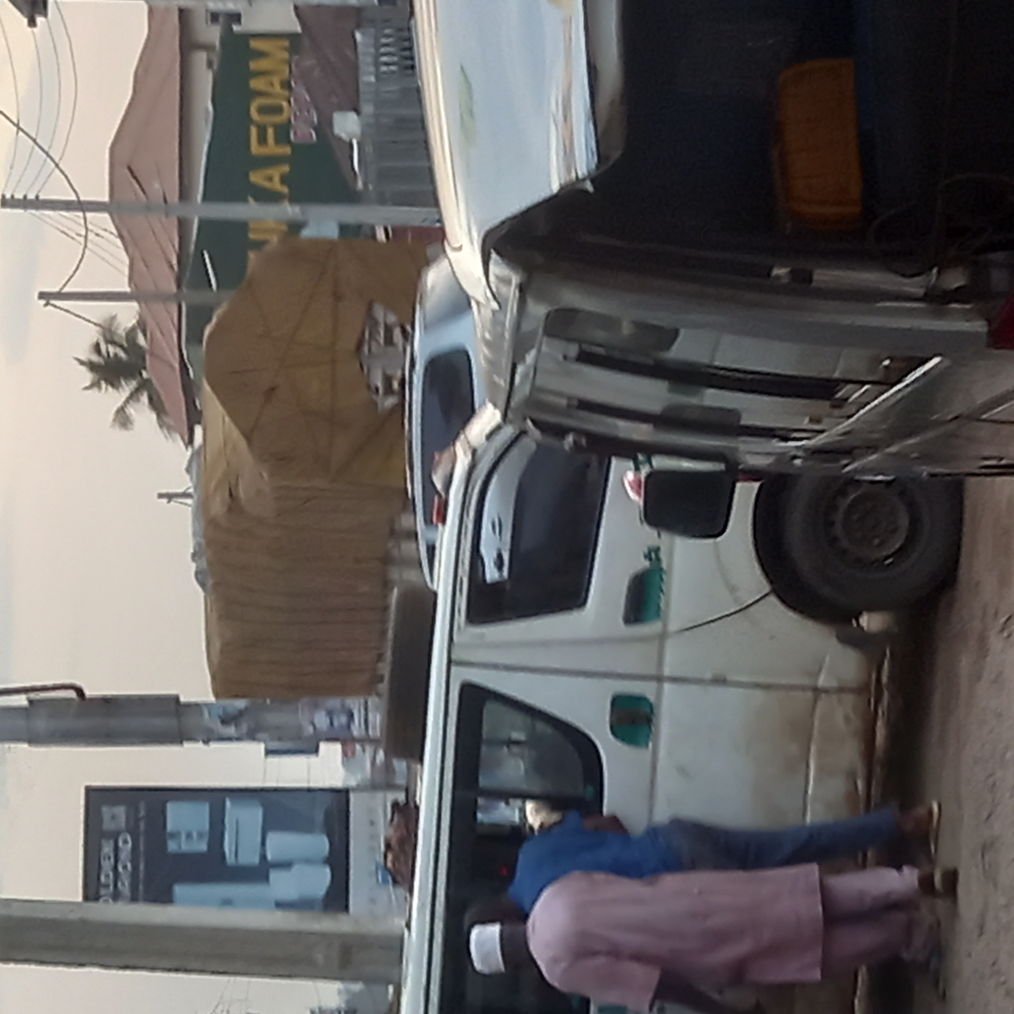 Two men stand next to a white van parked on a street in Dadi Riba, Nigeria. One man wears a pink shirt and white cap; the other, a blue shirt and pants. The van is loaded with goods covered with a brown tarp. A sign for "Foam" is visible in the background. A billboard showing bathroom fixtures stands on a pole.