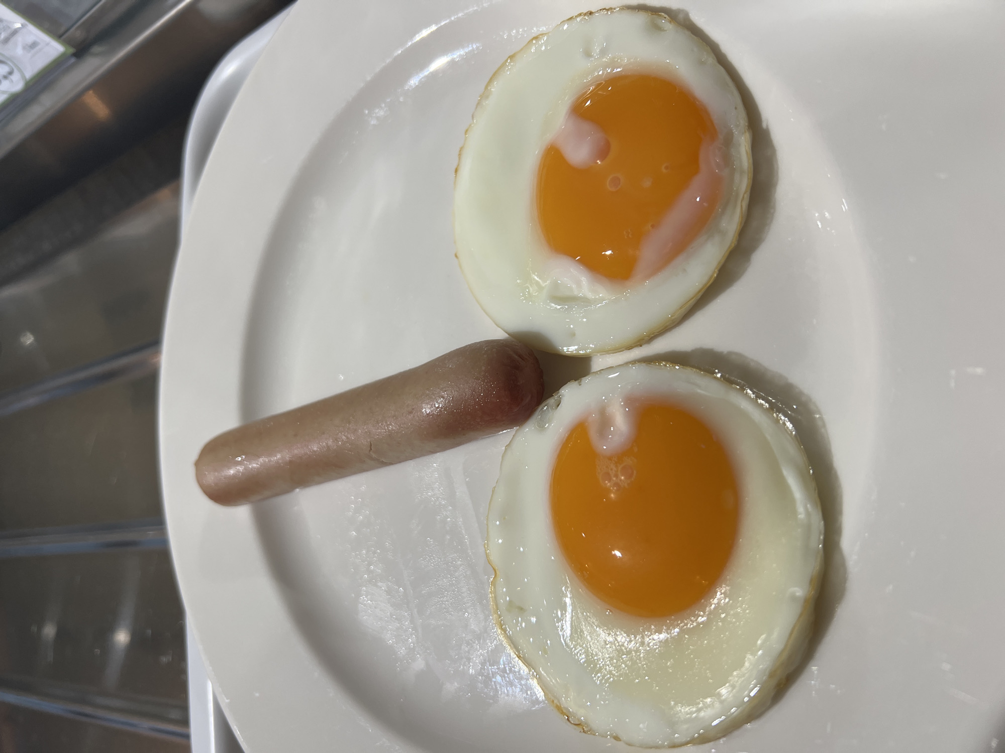 A close-up, eye-level shot captures a simple meal consisting of two fried eggs and one sausage served on a white, slightly oval plate. The two eggs are sunny-side up, featuring bright orange yolks that appear slightly runny or moist, surrounded by firm, white albumen with slightly crispy, golden-brown edges. Between the two eggs lies a single, light brown sausage, which also shows a slight sheen on its surface.
The white plate occupies the majority of the frame, presented against a blurred background of what appears to be a metallic kitchen or dining area surface, possibly a sink or countertop, with horizontal elements suggesting a rack or grooved design. The lighting is soft and even, highlighting the textures of the food. There are no people visible, and no specific time of day or weather can be discerned from the image, although the meal is typical of breakfast. No discernible text is visible on the plate or food items, though there's a very faint, illegible white label or marking in the blurred background. The image provides no visual cues to confirm the specified location of Budva, Montenegro.