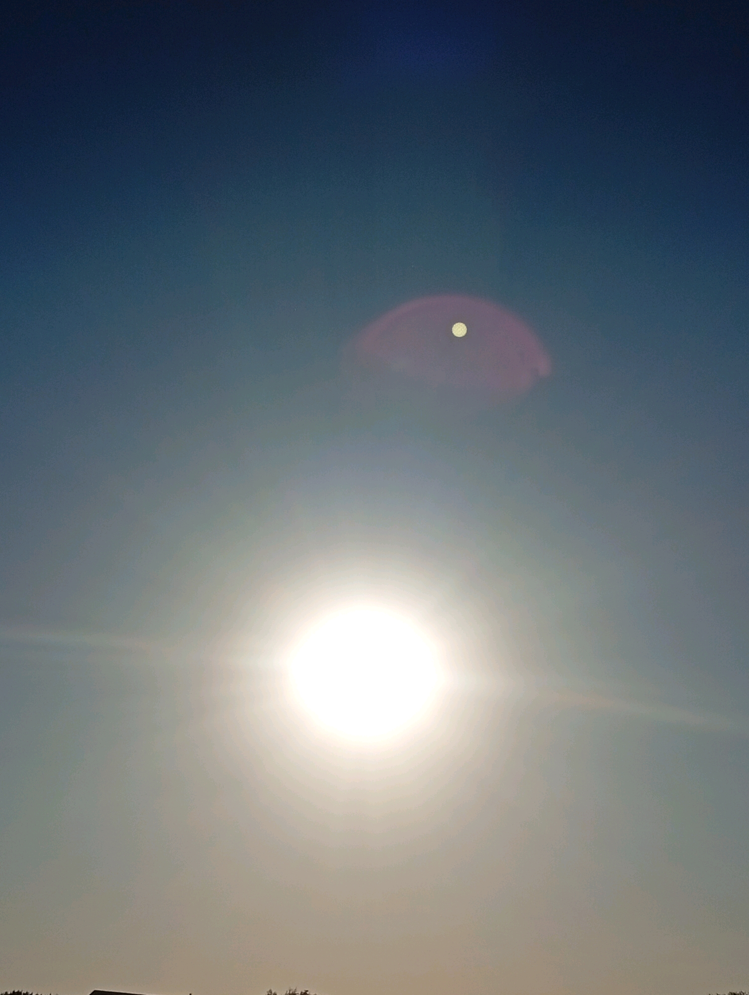 The image is a low-angle, outdoor shot of the sky with the sun prominently featured. The sun is a bright, white sphere in the lower-center of the frame, emitting strong lens flares that extend horizontally across the image. Above and slightly to the left of the sun, a smaller, white circular object is visible within a faint, pinkish-purple oval lens flare. The sky transitions from a lighter blue near the horizon to a deep blue at the top. The bottom edge of the image shows a dark silhouette of trees and what appears to be a building or structure against the bright sky.