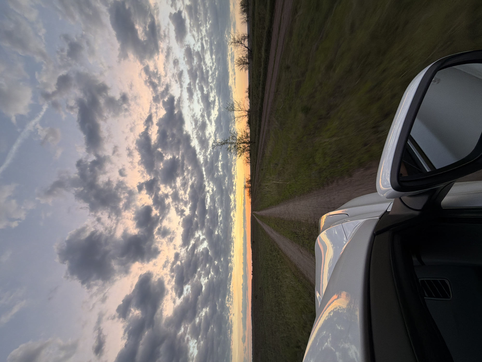 The image depicts a view from inside a moving vehicle. A white car's side mirror and a portion of the car's body are visible in the foreground. The background shows a dirt road stretching into the distance, flanked by grassy fields. The horizon line is marked by sparse trees and a vibrant sunset, transitioning from orange to blue. The sky above is filled with clouds. The location might be in the area of Nizhnyaya Lipovka, Russia, considering the rural landscape with a dirt road and horizon trees.