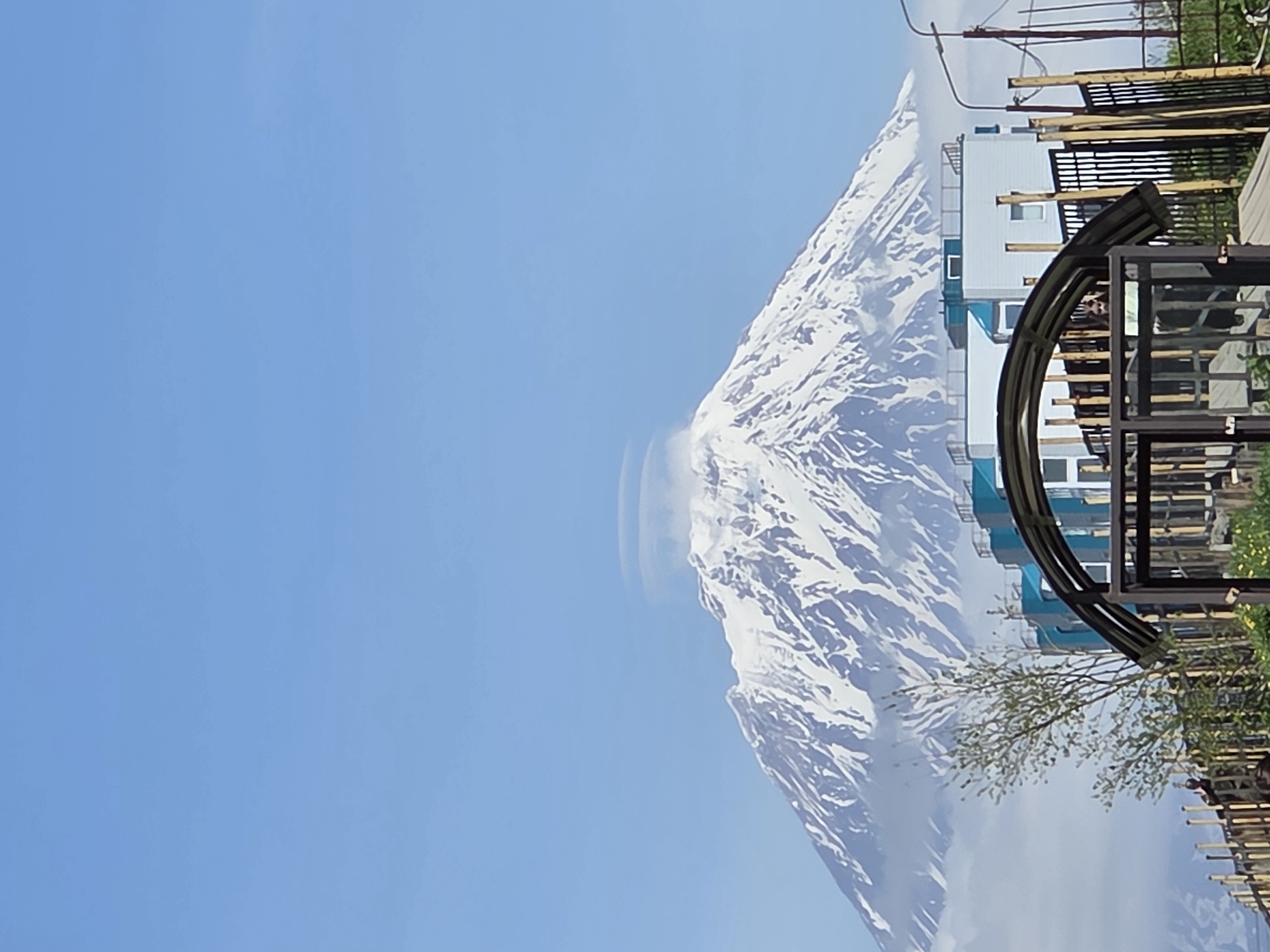 An imposing snow-capped mountain dominates the background under a clear blue sky. A lenticular cloud hovers over its summit, and mist clings to its lower slopes. In the foreground, a modern apartment building with teal accents stands behind a dark metal fence and archway. A single, slender tree with sparse green leaves is situated between the fence and the building. The scene appears to be a scenic view from an urban area, likely a residential development, looking towards a natural landmark.