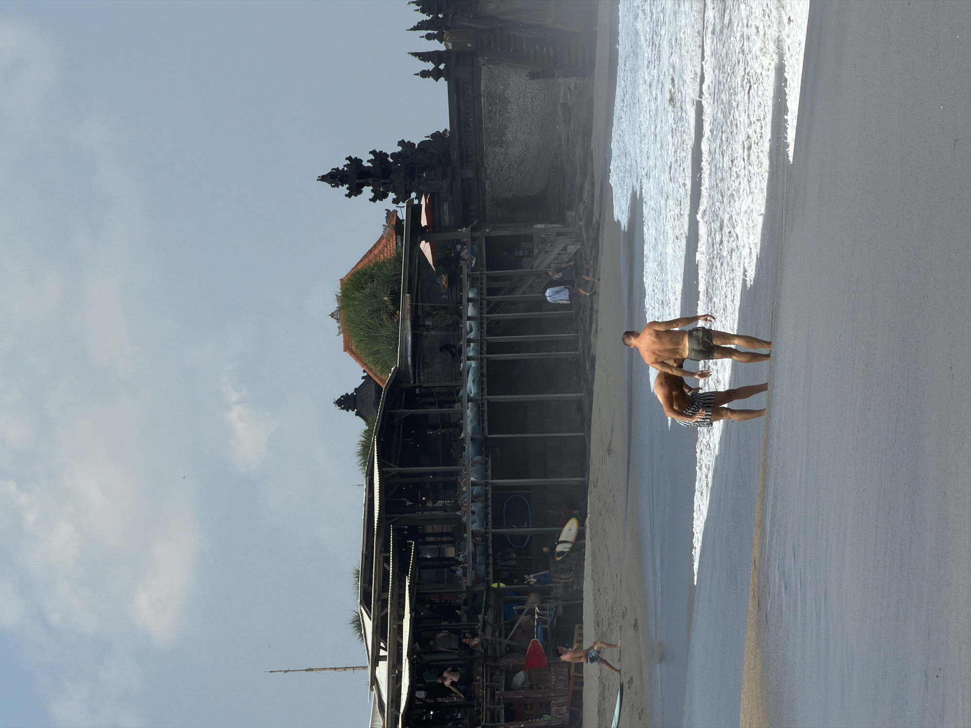 This is a vertical, medium shot of a beach scene in North Kuta, Indonesia. In the foreground, two men are walking near the water's edge. The man on the left is wearing black and white striped shorts, and is bent over, looking down. The man on the right is wearing green shorts and looking ahead. In the midground, there is a large, multi-level structure with a thatched roof, possibly a restaurant or bar, built on stilts over the sand. Several people are visible on the platforms and balconies of this structure. A dark stone temple structure with tiered roofs and intricate carvings is visible on the right side of the frame, behind the wooden building. The beach has dark sand, and the ocean in the foreground has white waves. The sky is overcast with a few clouds.