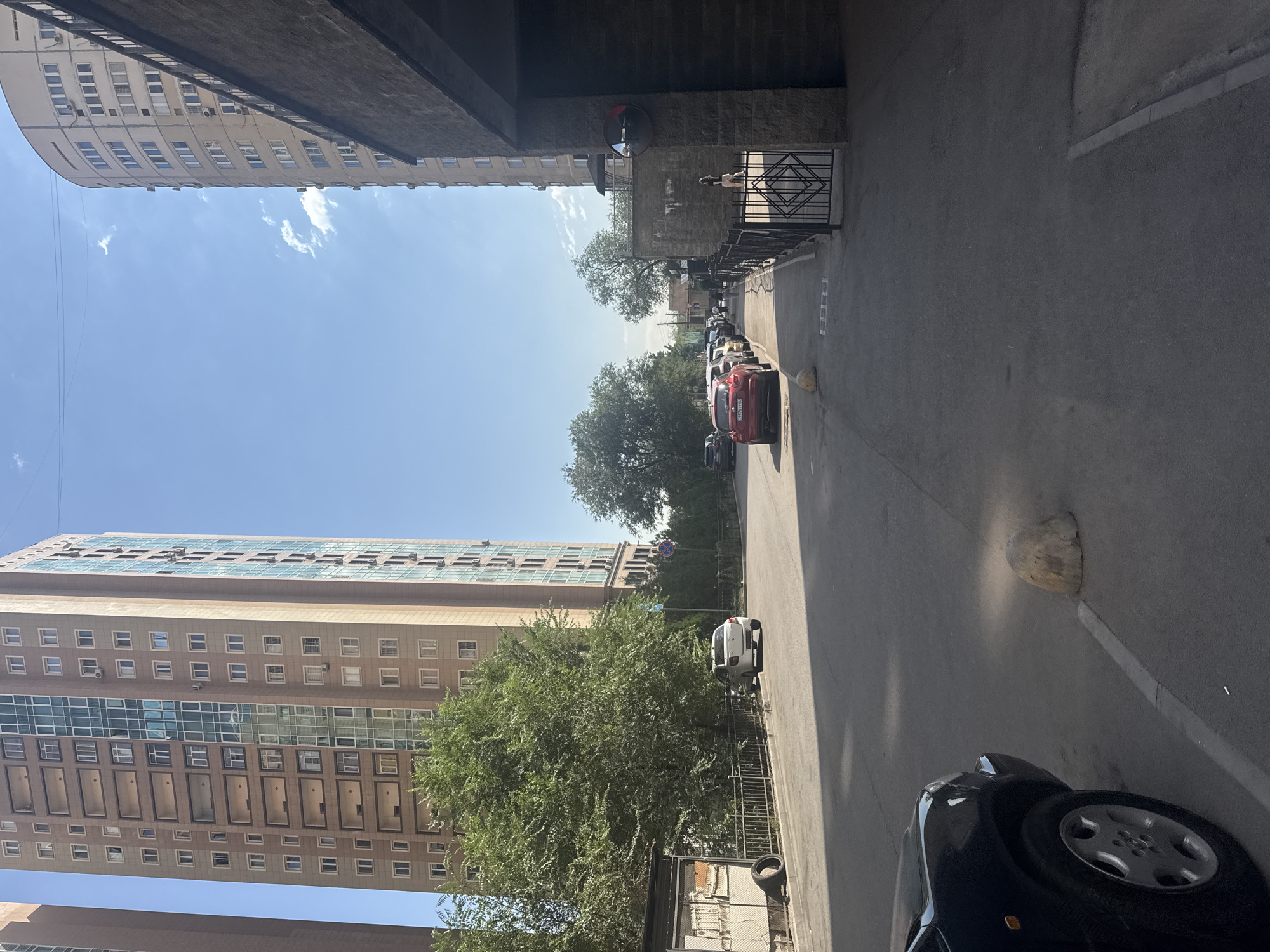 This image captures a street scene in Almaty, Kazakhstan, likely on a sunny day given the clear blue sky with scattered clouds. Two tall apartment buildings dominate the background. The building on the left is a modern, multi-story structure with a light brown facade and numerous windows arranged in a grid pattern. It also features balconies and a vertical section of glass windows. The building on the right is also tall and appears to be cylindrical in shape, with a pale beige exterior and many windows. A dark, textured building, possibly a parking structure or an underpass, is in the foreground on the right, casting a shadow onto the street.
The street itself is paved with asphalt and appears to be a residential or mixed-use area. Several cars are parked along the street and driving on it. A white car is parked on the left side of the road near a low fence. Further down the street, several other cars are parked, including a prominent red car. Trees line parts of the street, with a large, dense green tree on the left providing shade. A black car is partially visible in the bottom left corner of the frame. In the foreground, a large, light-colored concrete bollard is situated on the pavement. A metal fence with a geometric pattern is visible along the right side of the street, bordering the dark building. A convex mirror is mounted on the wall of the dark building, reflecting a portion of the street. The scene suggests a typical urban environment within Almaty.