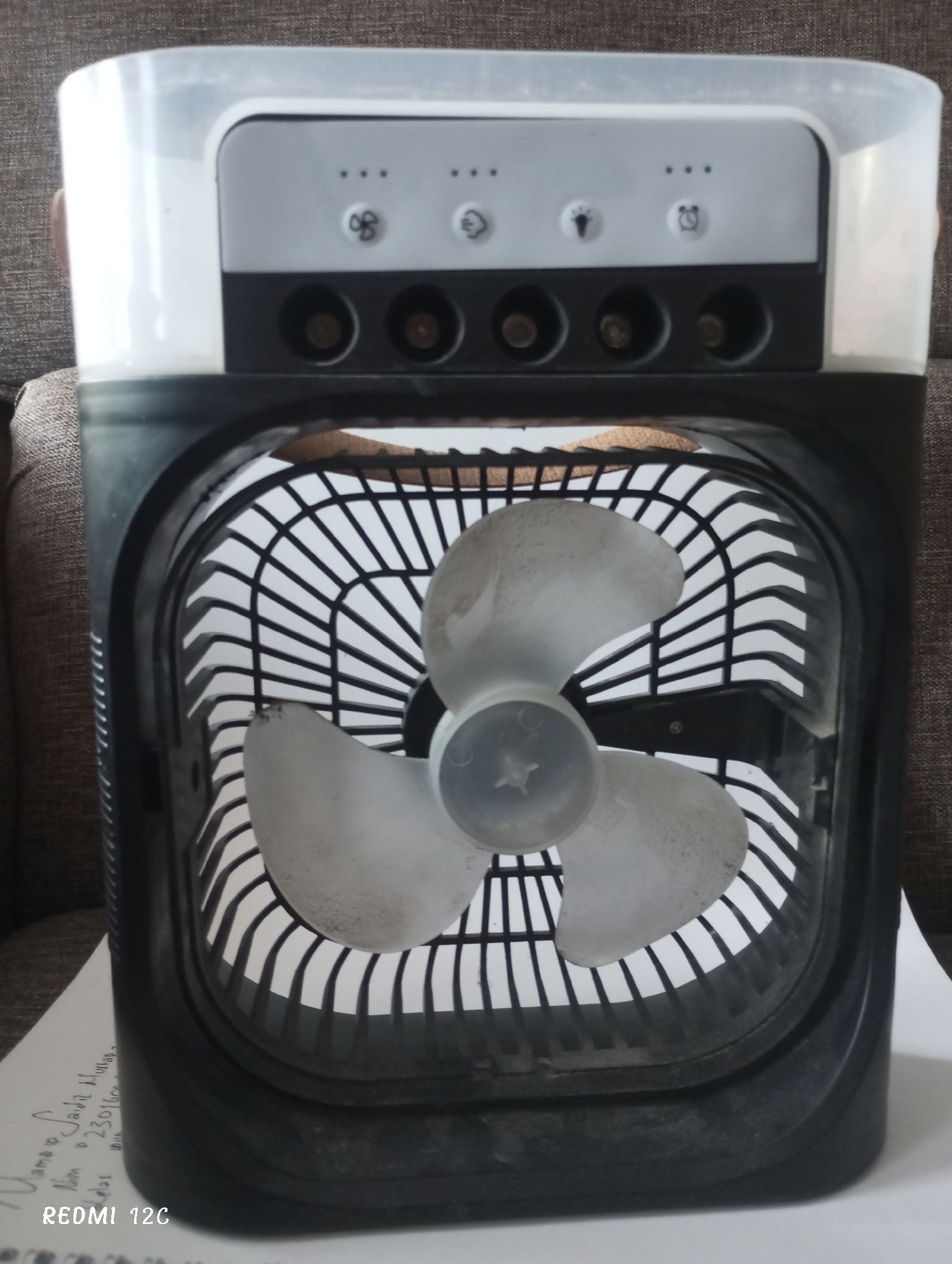 A close-up of a personal air cooler is centered in the frame. The cooler is black with a transparent white top that features control buttons. These buttons include symbols for speed, a water mist, a lightbulb, and a clock. There are corresponding cylindrical knobs directly below these buttons. The front of the cooler has a square grill to protect a three-bladed fan. The fan and grill appear slightly dusty. The cooler is resting on a white surface. Handwritten text is visible on this surface in the lower-left of the image. The location of Lhokseumawe, Indonesia is not identifiable from this image.