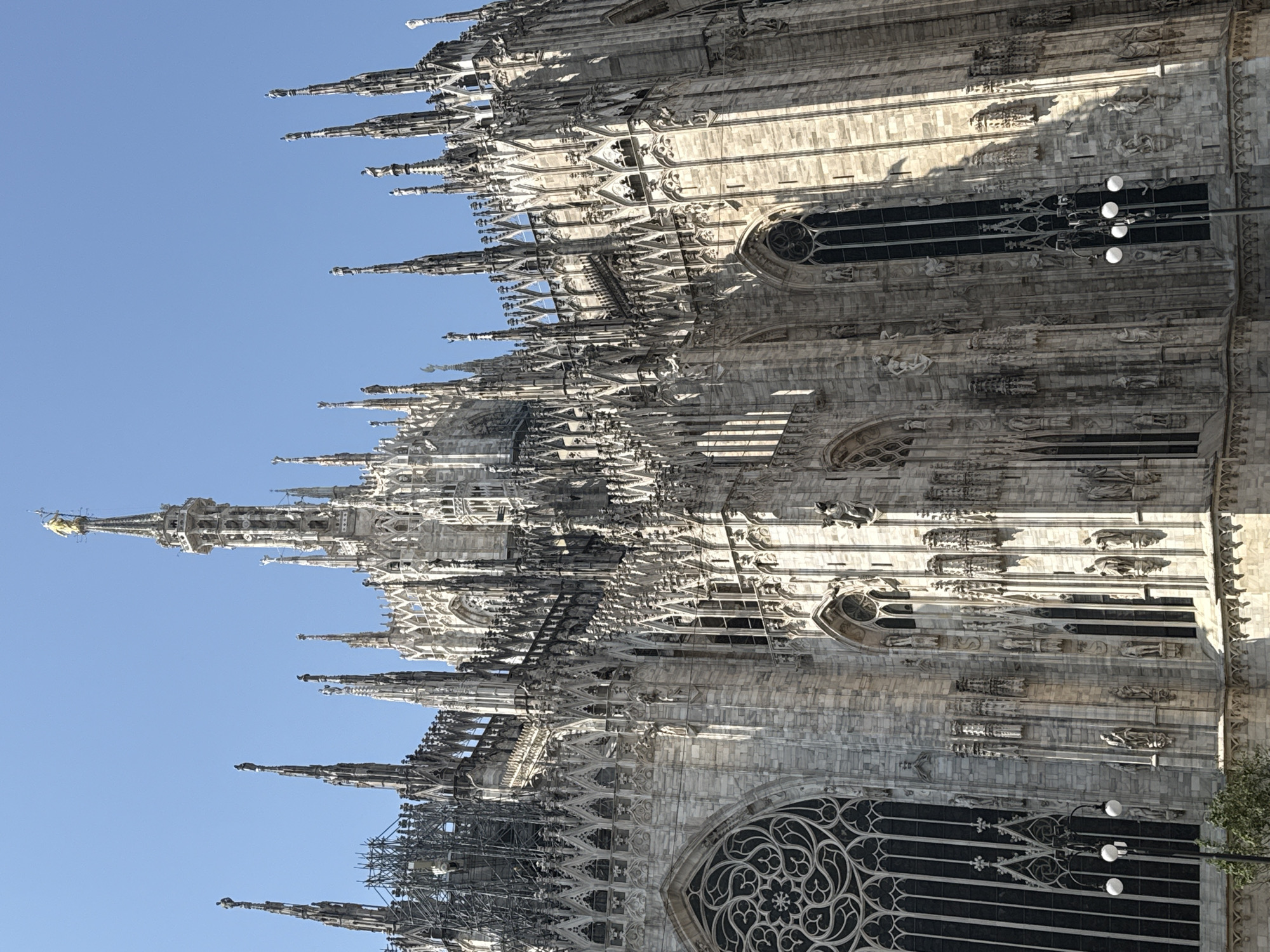 An upward-angled, close-up shot captures the intricate Gothic architecture of the Milan Cathedral (Duomo di Milano) against a clear blue sky. The massive structure is constructed from light-colored marble, featuring numerous ornate spires (pinnacles) that ascend towards the top of the frame. Many of these spires are adorned with detailed sculptures and delicate tracery.
The cathedral's facade is rich with decorative elements, including pointed arches, rose windows, carved statues, and relief work that showcase the intricate craftsmanship. Strong sunlight illuminates the marble, creating deep shadows that emphasize the three-dimensional depth of the carvings and architectural details.
At the apex of the tallest visible spire, a golden statue, identifiable as the Madonnina, gleams brightly against the sky. In some sections, particularly on the lower left and upper right, scaffolding is visible, indicating ongoing restoration or maintenance work. A large, dark-paned window with elaborate tracery is prominent in the lower right, and a street lamp with three white globes is partially visible on the right side. A small hint of green foliage from a tree is present at the bottom right corner. The sky is a uniform, cloudless blue, suggesting a clear day, likely morning or afternoon, given the direction of the sunlight. No people or visible text are present in the image.