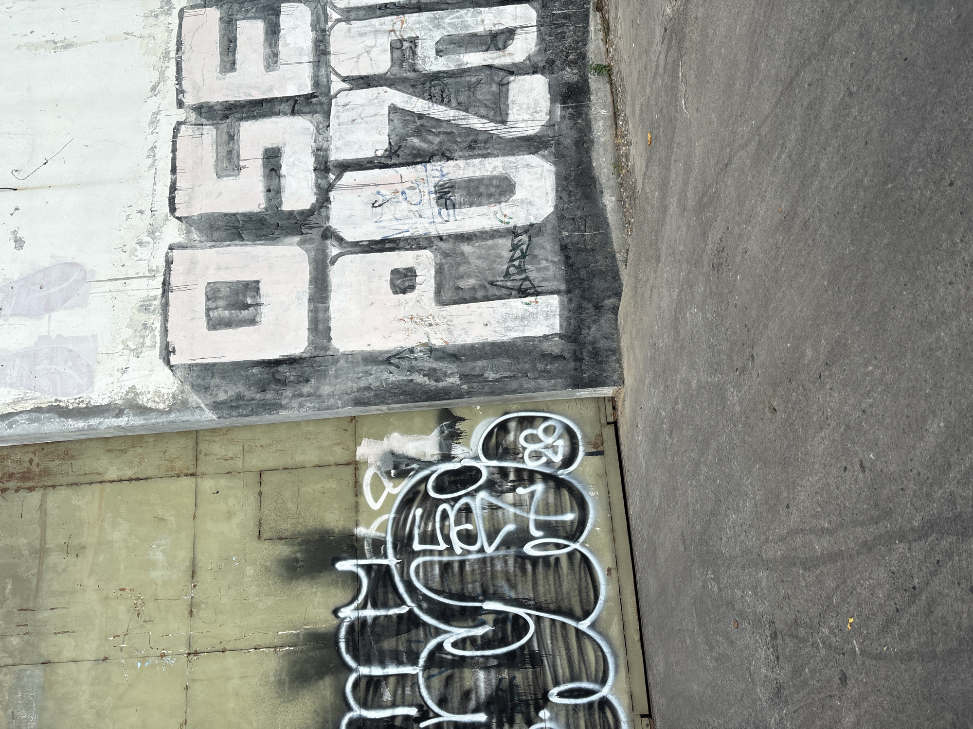 The image depicts a section of a concrete wall in Cirebon, Indonesia, covered in graffiti. On the left side, a large, light green metal panel, showing signs of rust and wear, is adorned with white bubble-style graffiti that appears to be a tag. The graffiti has black spray-painted shadows. To the right of the metal panel, a rough, light-colored concrete wall exhibits more elaborate graffiti in large, blocky white letters with black outlines and drips. The text reads "OSE PUZA". The concrete wall also shows patches of peeling paint and faint, older graffiti or marks underneath the main artwork. The ground in front of the wall is a gray asphalt surface, appearing somewhat weathered and showing some debris. The overall scene suggests an urban environment where public surfaces are used for artistic expression, possibly an underpass or a large outdoor wall.