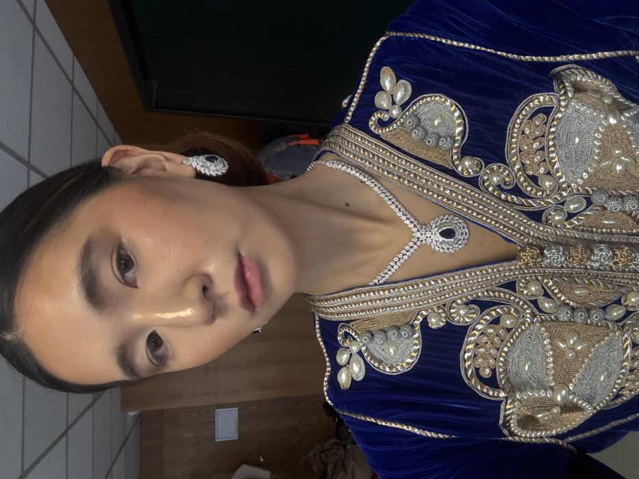 A close-up portrait captures an East Asian woman from the chest up, positioned indoors. Her dark hair is styled back, and she wears subtle facial makeup, including a highlight along the bridge of her nose. She is dressed in a royal blue velvet garment, likely a caftan or traditional dress, adorned with intricate gold, silver, and cream-colored embroidery, complemented by white pearls and beads on the collar and upper torso. She wears a matching high-jewelry set: a necklace featuring a large tear-drop shaped dark blue gemstone pendant, encircled by numerous pavé-set clear gemstones, with a similar clear gemstone chain; and corresponding tear-drop shaped earrings, each with a dark blue gemstone surrounded by clear pavé-set gemstones arranged in a stylized, elongated pattern. The background consists of a plain light-colored wall with visible ceiling tiles on the left side of the frame, and a dark wooden panel or door on the right. No identifiable landmarks or specific architectural features are present to determine a precise location within Paris, France; the environment appears to be a generic interior room, possibly a dressing or preparation area.