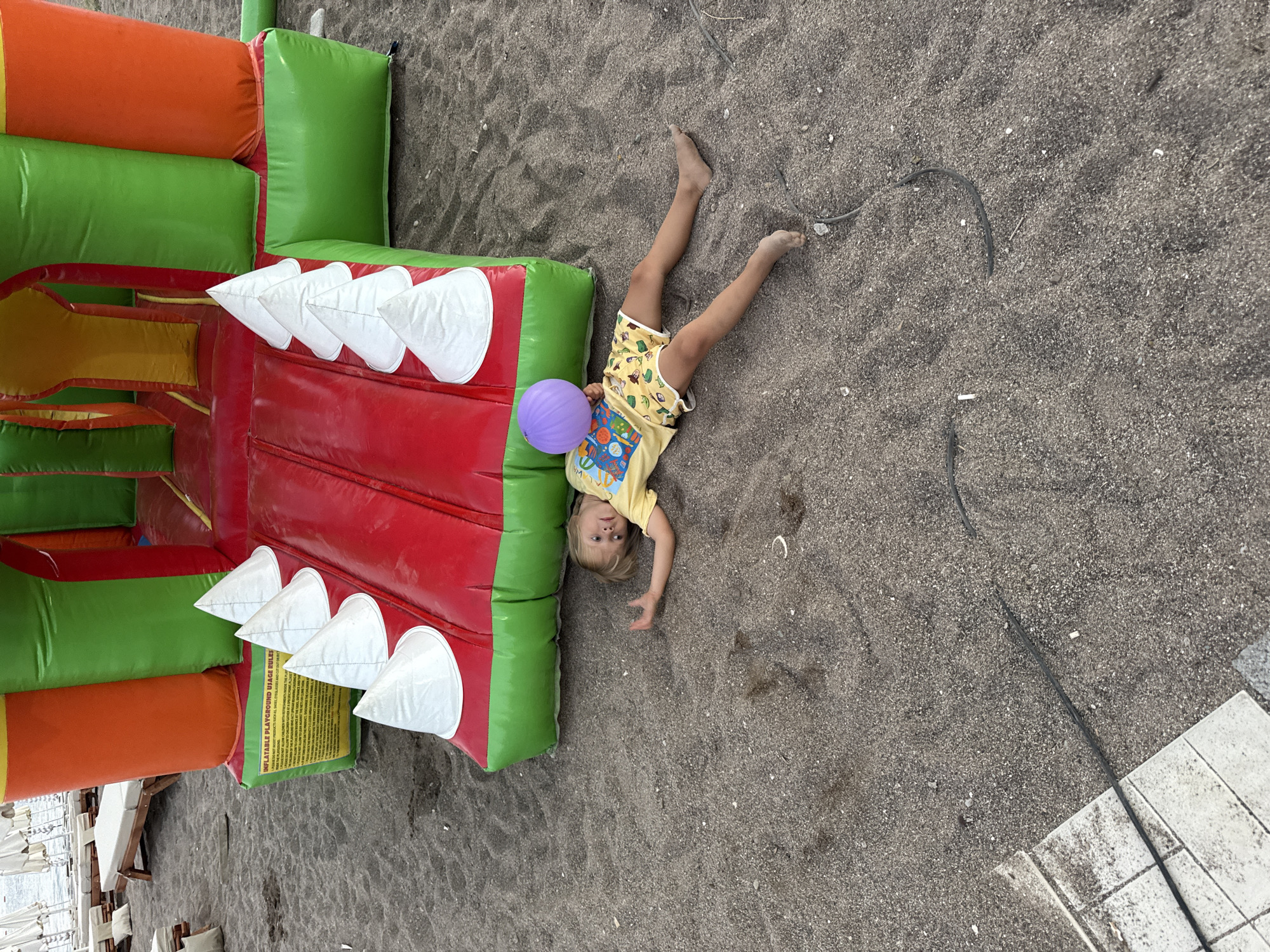 A blonde-haired child lies on their back on a dark sand beach in Bečići, Montenegro, next to a large, colorful inflatable playground. The child is wearing a yellow t-shirt with a graphic print and yellow shorts with a pattern. They are holding a purple ball in their right hand, which is partially obscuring their face. The inflatable playground is red, green, and orange and features several white conical protrusions along its lower edge. In the background, beach chairs and umbrellas are visible near the water. The sand has some debris, including black cords and small white objects.