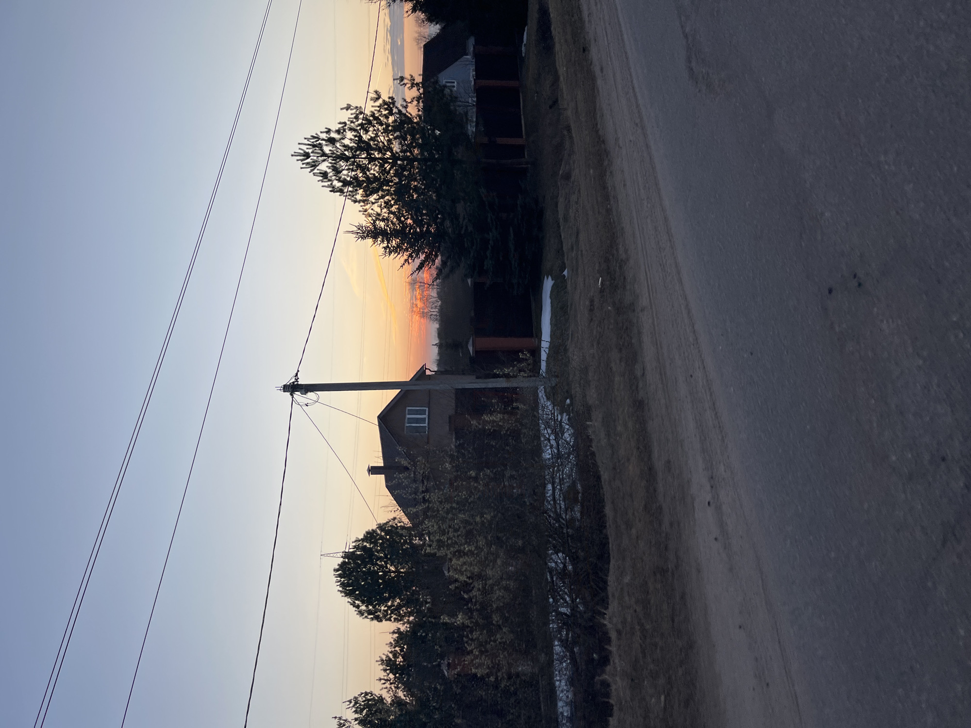 The image shows a street scene in Раздолье, Russia at sunset. The sky transitions from blue in the upper left to orange on the horizon. A road runs along the right side of the image. On the left side of the road, there are houses and trees. Power lines are visible stretching across the sky. A utility pole stands near one of the houses. Patches of snow remain on the ground near the houses. The scene appears quiet and still. There are no visible people, actions, or interactions.