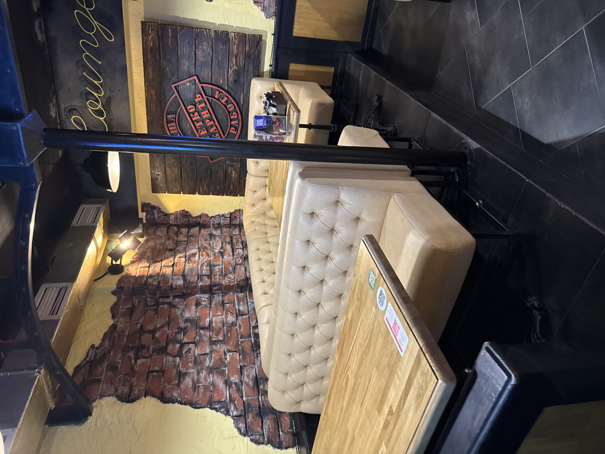 The image displays the interior of what appears to be a lounge or cafe, oriented vertically. The space features a rustic industrial aesthetic combined with comfortable seating.
In the foreground and mid-ground, there are multiple sections of light beige, tufted booth seating. Adjacent to these booths are light-colored wooden tables, one of which has stickers including a QR code and a label with small red text. A prominent black metal post or railing runs horizontally across the mid-section of the image.
The walls exhibit varied textures. One wall features exposed red brickwork with areas of yellow, distressed plaster. Another section of the wall behind the booths is covered with dark wooden planks, above which is a sign with partially visible Cyrillic text in red and black. Above the main seating area, a dark sign with the word "Lounge" written in yellow cursive script is visible. The floor is covered with dark, possibly tiled, material.
Artificial lighting illuminates the scene, with spotlights visible on the brick wall and a diffused light source hanging near the "Lounge" sign, suggesting an indoor setting with low natural light, likely in the evening. No people or specific activities are depicted in the static shot. The Cyrillic text on the signs indicates a Russian-speaking location.