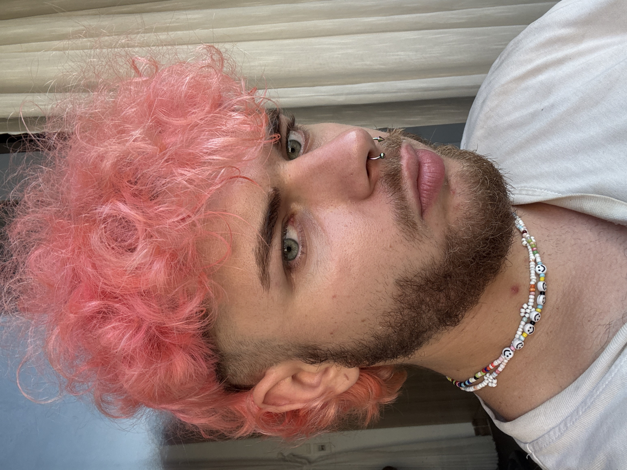 A close-up, tilted portrait shows a man with pink curly hair. He has a beard and fair skin, green eyes, and multiple piercings in his nose. He's wearing a white t-shirt and a beaded necklace with smiley face charms. The background is a neutral-colored surface. There is no indication of the location being Đà Nẵng, Vietnam.