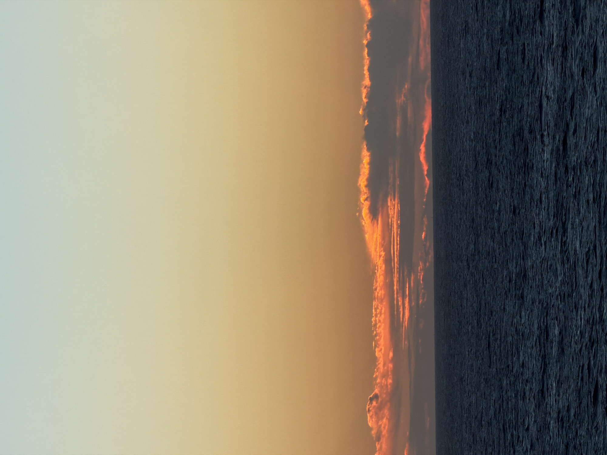 The image depicts a sunset over water. The sky transitions from a pale blue to a warm orange. A band of clouds with orange and red highlights is visible on the horizon line, marking the boundary between the sky and the water. The water appears dark and textured, suggesting waves or ripples. The location is unspecified.