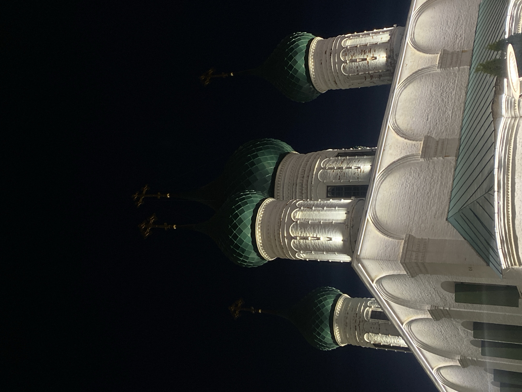 At night, the image depicts an illuminated Orthodox church, likely located in Russia, given its architectural style. The building features a pale facade, accented by arched details and pillars. Three domes with green, patterned exteriors are visible, each topped with a golden Orthodox cross. The structures are brightly lit against a stark black sky.
