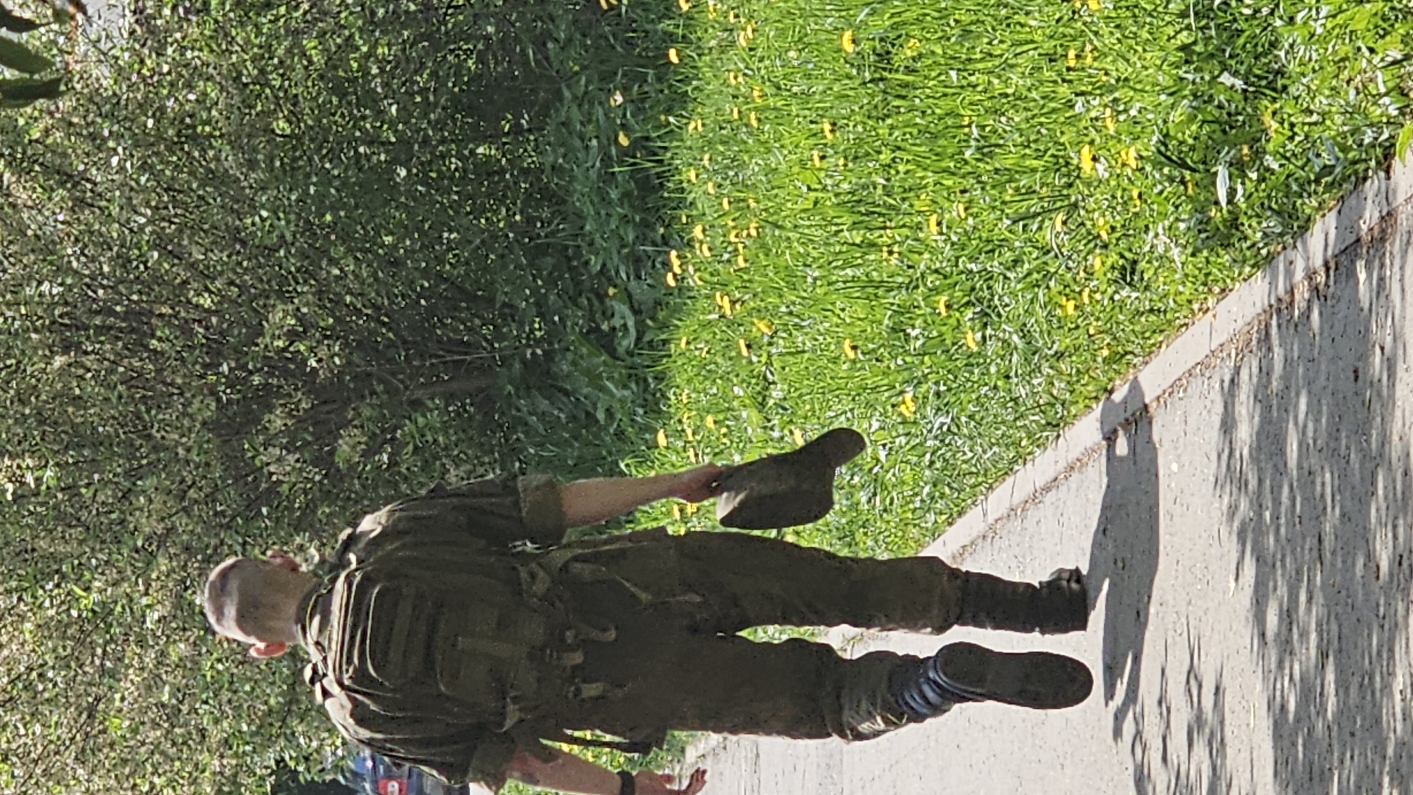 A full-body shot captures a soldier in camouflage gear walking on a sidewalk next to a grassy area dotted with yellow flowers. The soldier, seen from the back, carries a hat in one hand. The sidewalk is concrete and displays shadows from a tree. Lush green foliage extends from the left side of the frame. The city of location is unknown, as is its province.