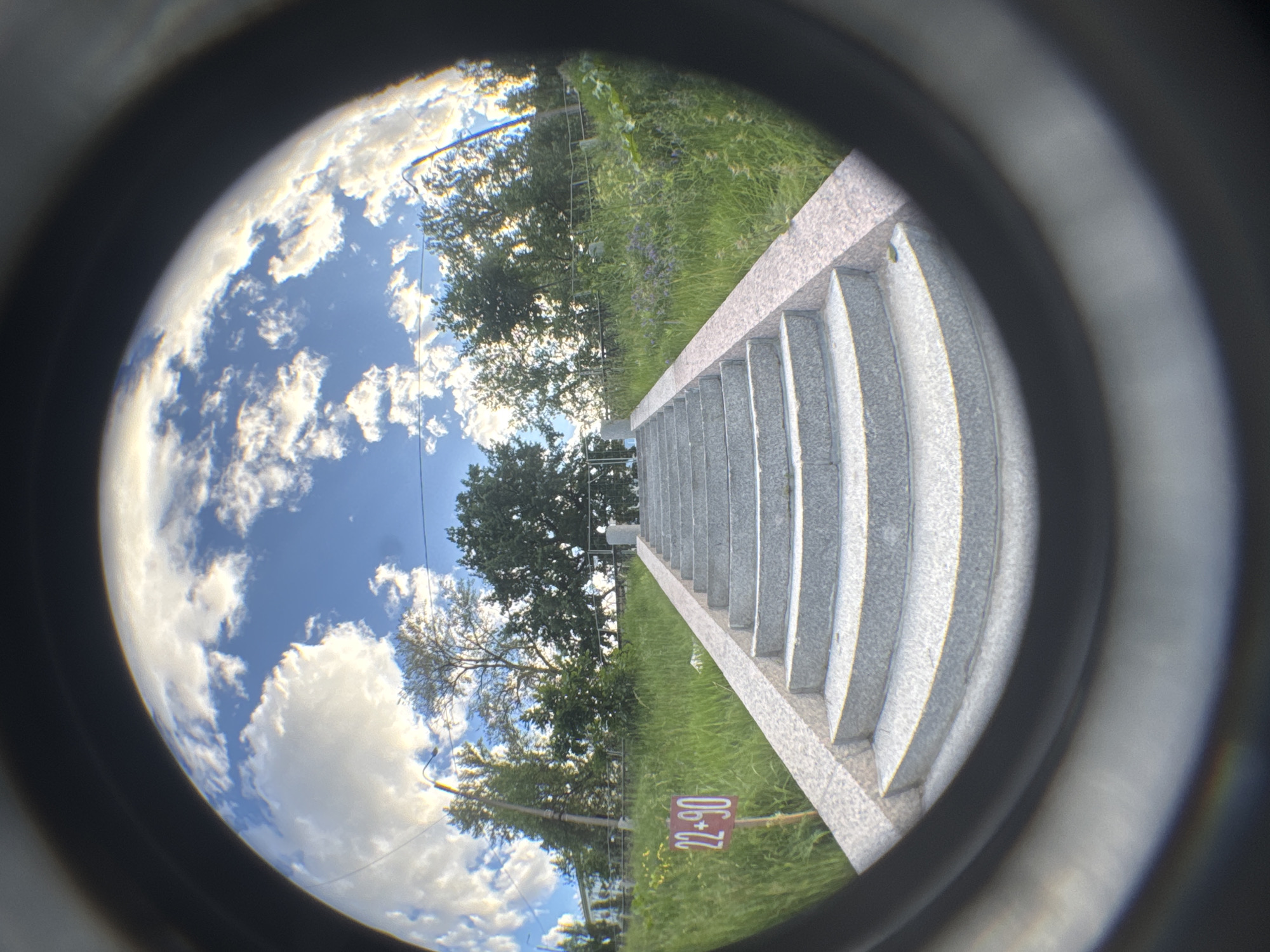 This is a fisheye lens photograph taken on a sunny day at an outdoor staircase in Saint Petersburg, Russia. The staircase is made of granite and ascends towards a grassy slope dotted with trees and vegetation. A red sign with the numbers "22+90" is visible to the left of the staircase. The sky above is blue with scattered white clouds, and a faint rainbow effect is present at the bottom edge of the image due to the lens distortion. There are no people visible in the scene. The overall impression is one of a serene, urban park setting.