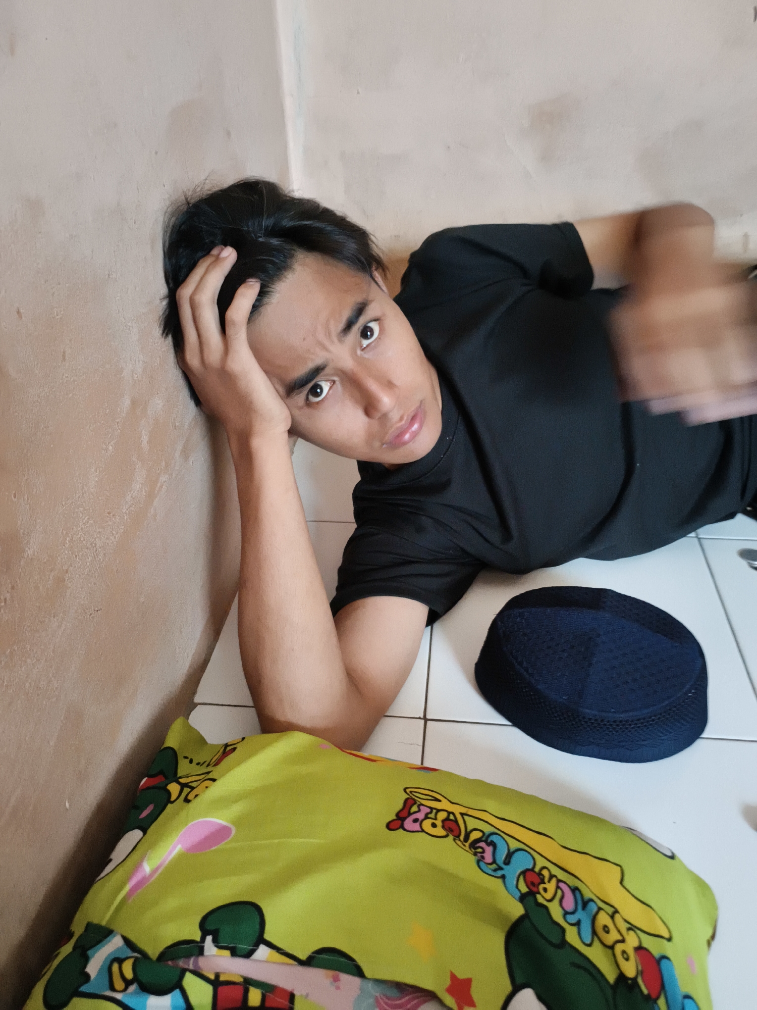 A young man lies on a white tiled floor near a beige wall, resting his head in his hand. He is wearing a black t-shirt. Next to him is a dark blue knitted skullcap (kufi). Beneath him is a yellow pillow or cushion with cartoon-like designs. The location is in Cianjur, Indonesia.