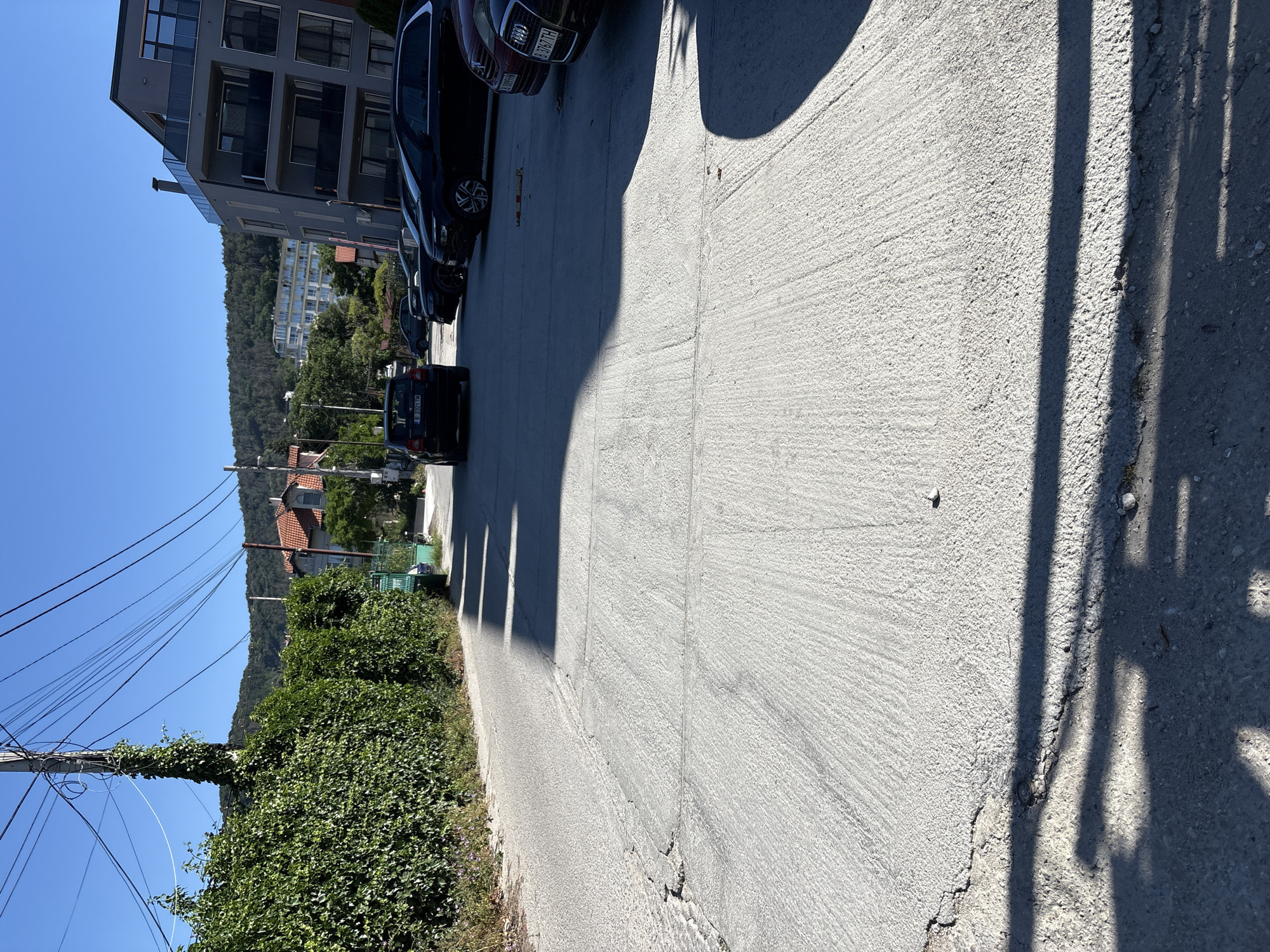 The image depicts a paved area, likely a street or parking lot, in Varna, Bulgaria. The ground is a light gray concrete, showing signs of wear and cracking, particularly along the left side. In the background, a dense green hedge lines the left side of the paved area, partially obscured by shadows cast by an unseen structure. To the right of the hedge, a utility pole covered in ivy stands tall, draped with numerous electrical wires against a clear blue sky.
Further in the background, several cars are parked. A black hatchback with license plate "B 2321 TM" is visible on the left. To its right, a line of black sedans and SUVs occupy parking spaces. A modern gray building with multiple balconies and windows rises on the far right side of the frame. Hills covered in trees are visible in the distance, under the bright blue sky. The scene appears to be an outdoor area with a mix of natural elements (hedge, trees) and urban infrastructure (utility pole, cars, building). The lighting suggests it is daytime, with strong sunlight casting distinct shadows across the paved surface.