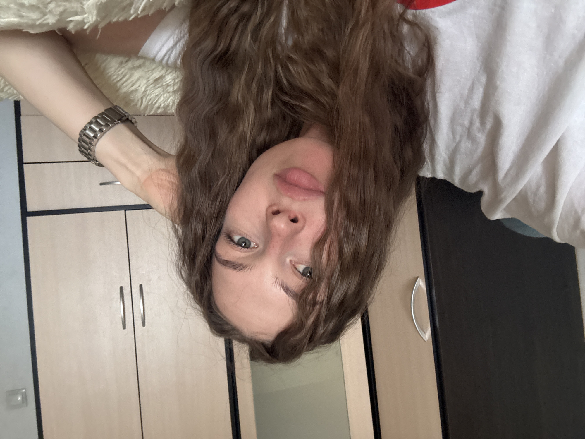 A young woman with long, wavy brown hair lies on her side, propping her head with her right hand, which is adorned with a metal wristwatch. She wears a white t-shirt. Her eyes are open and looking slightly to the left. The background features a closet with light-colored doors and dark trim. The scene appears to be indoors.