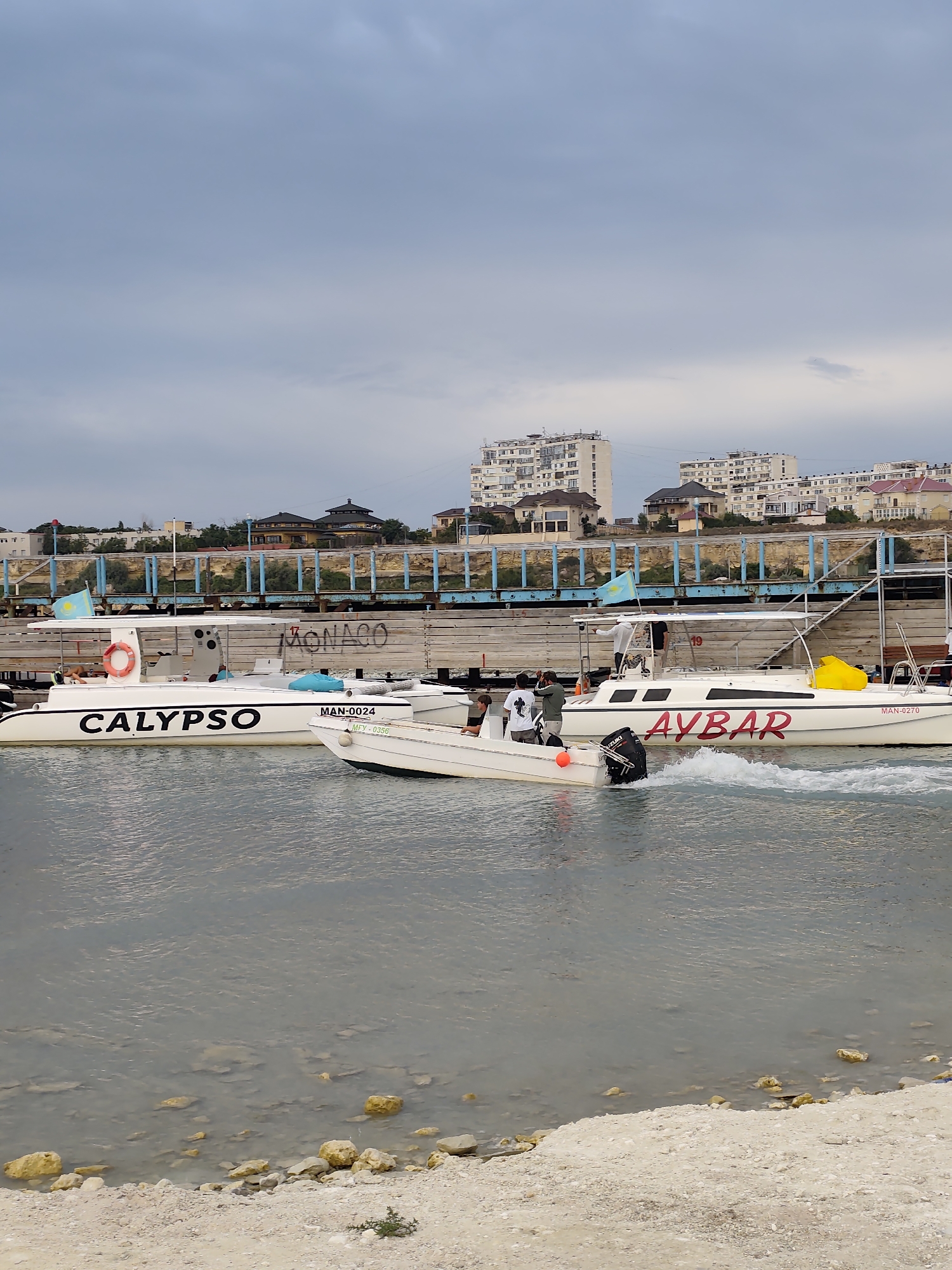 In Aktau, Kazakhstan, on a cloudy day, two white speedboats are docked near a wooden pier. The larger boat, named "CALYPSO" in bold black lettering, displays a "MAN-0024" registration number and a flag of Kazakhstan on its canopy. Beside it, another white speedboat, "AYBAR", also has a "MAN-0270" registration. In the foreground, a smaller white motorboat is in motion, creating ripples in the murky water, with three individuals visible onboard. The background reveals a waterfront promenade with buildings and a cityscape. The shore is covered with sand and rocks.