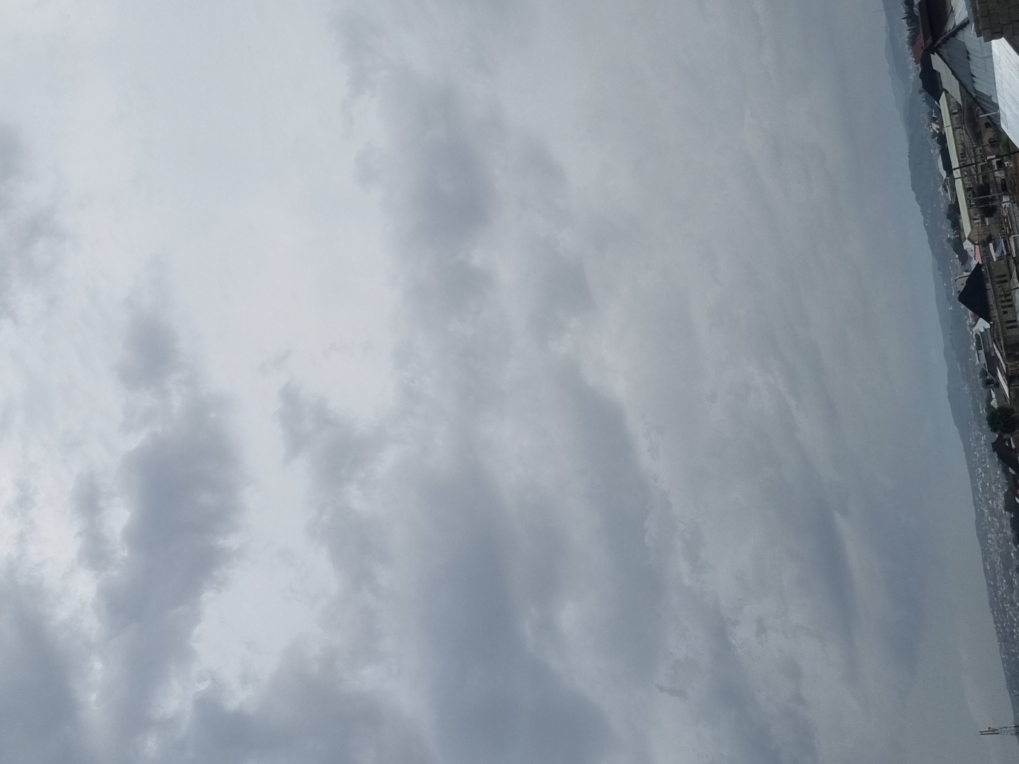 The image shows a cloudy sky over a city in Gwong, Nigeria. The clouds are dark gray and appear heavy, suggesting an impending storm or overcast weather. Below the sky, a city skyline is visible, characterized by numerous buildings with various roof types, including corrugated metal and what appear to be some tiled roofs. The buildings are densely packed, indicating an urban or suburban area. In the distance, rolling hills or mountains can be seen through the hazy atmosphere. The overall lighting is dim due to the cloud cover, giving the scene a somber mood. There are no visible people or specific actions occurring in the image; it is a static view of the environment. A tall, slender tower, possibly a communication or utility pole, is visible on the left side of the frame.