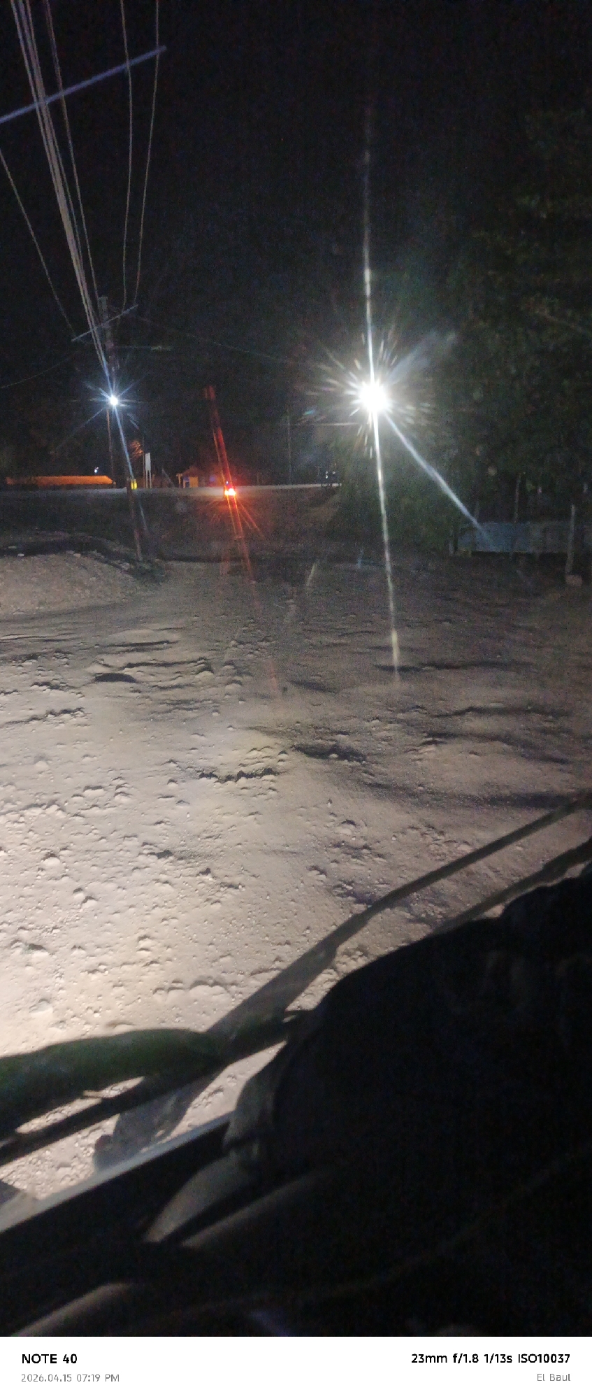 Captured from within a vehicle in El Baúl, Venezuela, this nighttime scene depicts a rural road bathed in the glare of streetlights. The road’s surface, uneven and textured, reflects the intense light, obscuring finer details. In the background, a faint building and a red light suggest the presence of structures along the road. Utility poles and power lines stretch into the night sky. The vehicle’s dashboard and windshield wipers are visible in the foreground, casting shadows and adding depth to the composition. The environment is dark, with the only illumination stemming from the streetlights.