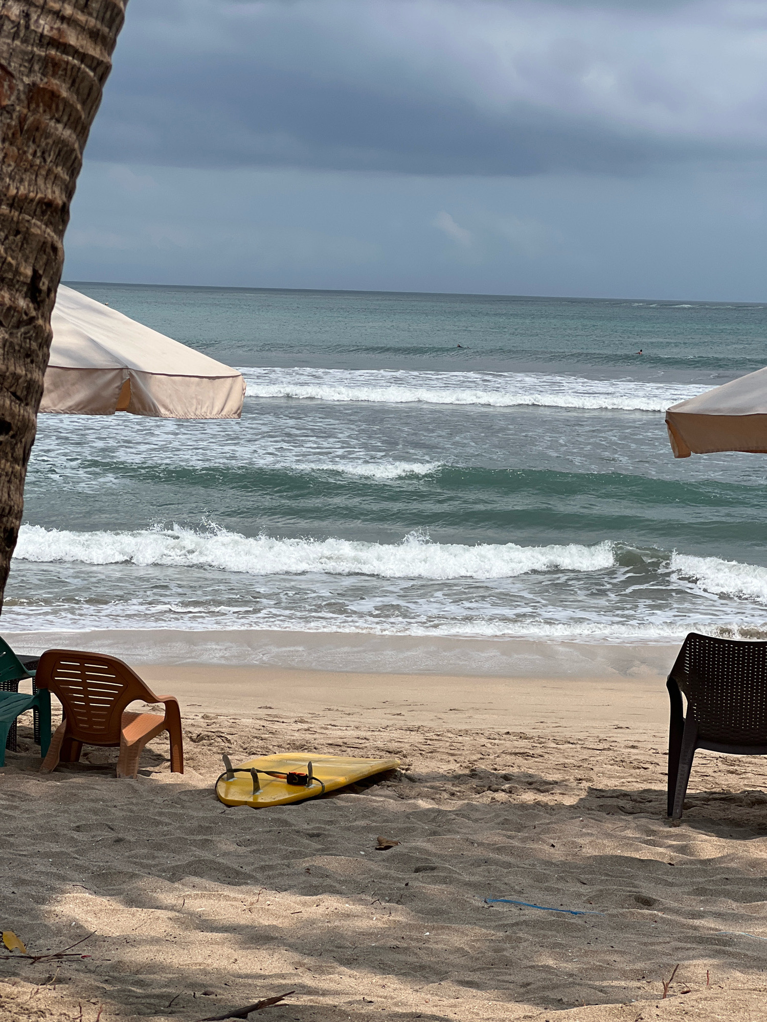 The image captures a serene beach scene in Kuta, Indonesia, under a dramatic overcast sky. In the foreground, the sandy beach is dotted with fallen leaves and branches, with a weathered palm tree trunk on the left providing partial framing. Two beach chairs are visible; one is a light brown plastic chair positioned to the left, and another is a dark brown, perforated chair on the right. A yellow surfboard with fins lies horizontally on the sand between the chairs. In the middle ground, gentle waves crash onto the shore, creating a foamy white surf. Two beach umbrellas, one on the left and one on the right, offer shade. The ocean stretches to the horizon, where it meets the gray, cloudy sky. A few distant figures can be seen in the water, suggesting the presence of surfers or swimmers. The overall atmosphere is one of calm and natural beauty, characteristic of Kuta's coastline.